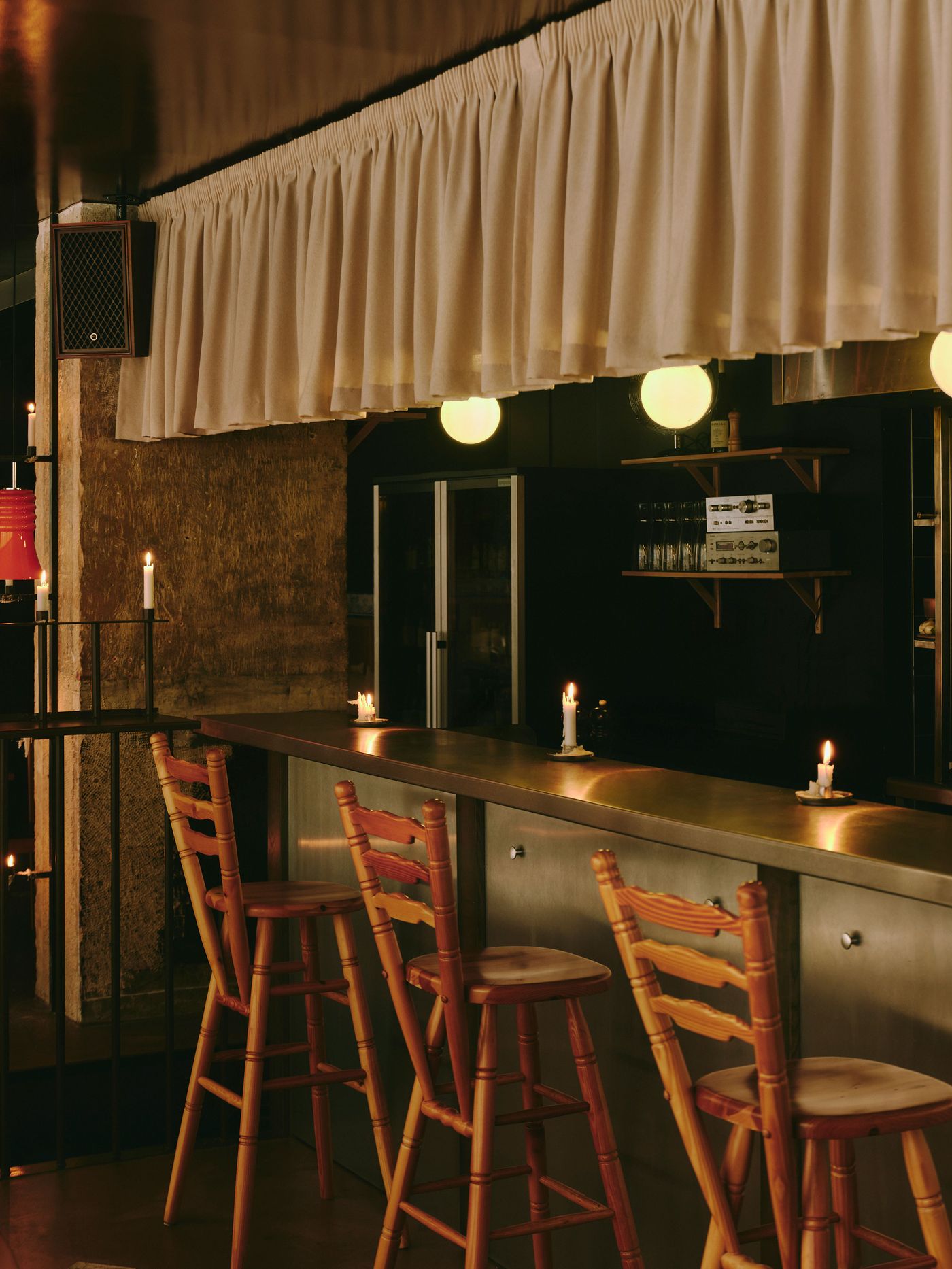 The bar at SaltoBar in Paris after dark, where turned-pine ladder-back stools pull up to a brushed stainless-steel counter dotted with candles. Above, a ruffled fabric valance hangs over opal globe lights. Vintage hi-fi components rest on wall shelves, and a wood-cased speaker is mounted on a textured concrete pillar. The bar at SaltoBar in Paris after dark, where turned-pine ladder-back stools pull up to a brushed stainless-steel counter dotted with candles. Above, a ruffled fabric valance hangs over opal globe lights. Vintage hi-fi components rest on wall shelves, and a wood-cased speaker is mounted on a textured concrete pillar.
