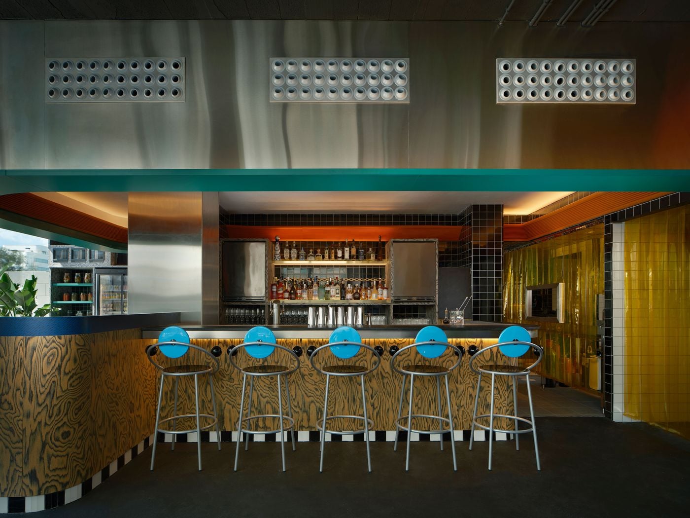 A symmetrical view of the bar at Seth & Manni's shows five First stools designed by Michele De Lucchi aligned before an ochre wood-grain counter edged in checkerboard tiles. Above, a brushed stainless-steel wall carries three perforated vent panels. An orange ribbed fascia and black tiles frame the illuminated back bar and an amber strip curtain. A symmetrical view of the bar at Seth & Manni's shows five First stools designed by Michele De Lucchi aligned before an ochre wood-grain counter edged in checkerboard tiles. Above, a brushed stainless-steel wall carries three perforated vent panels. An orange ribbed fascia and black tiles frame the illuminated back bar and an amber strip curtain.