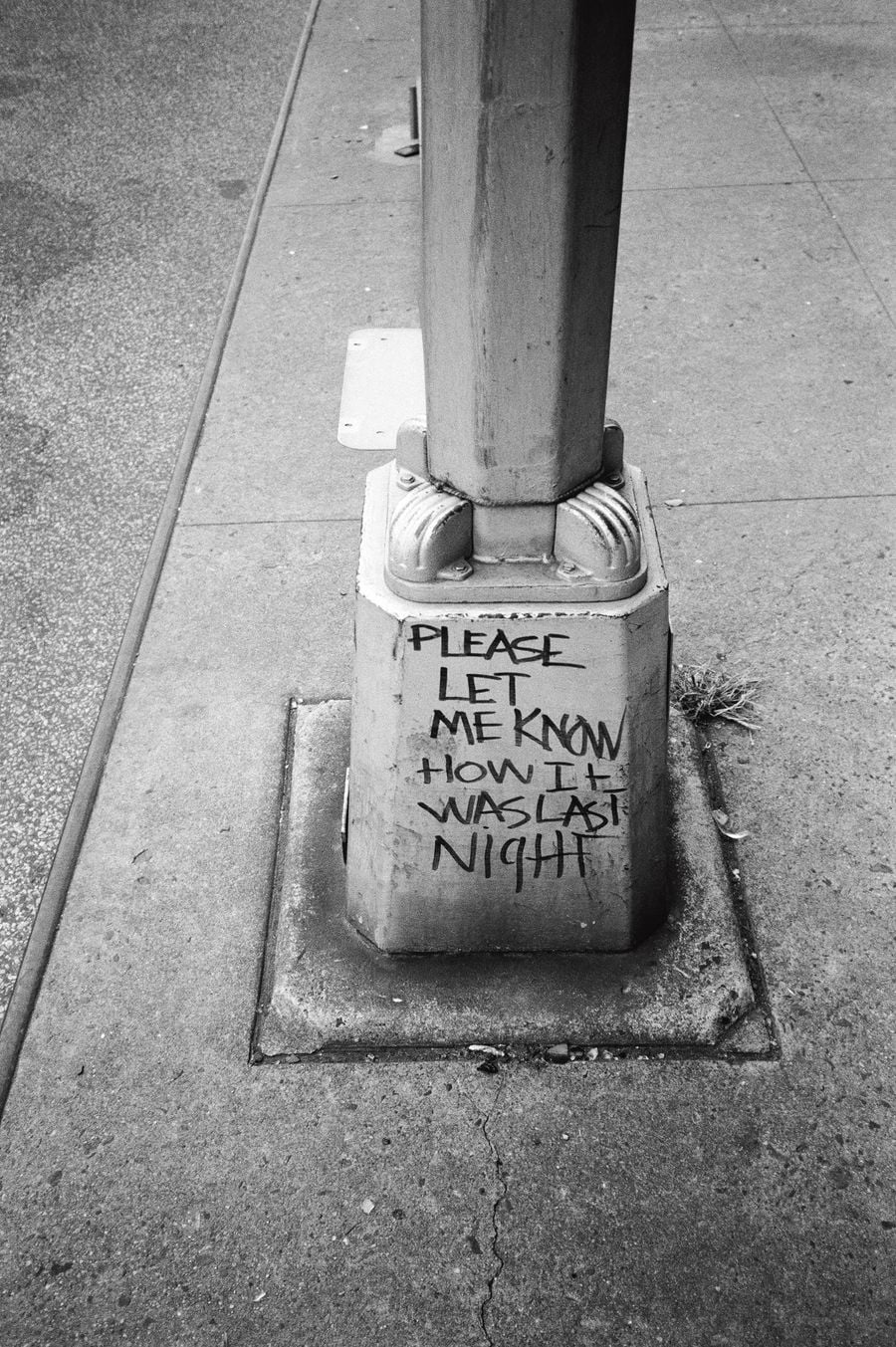 A black-and-white photograph by Stanley Stellar from Don't Worry Boys captures the base of a street lamp on a cracked New York pavement, scrawled with the handwritten plea "Please let me know how it was last night". A tuft of grass sprouts beside the concrete plinth, lending the anonymous message a wry, wistful intimacy. A black-and-white photograph by Stanley Stellar from Don't Worry Boys captures the base of a street lamp on a cracked New York pavement, scrawled with the handwritten plea "Please let me know how it was last night". A tuft of grass sprouts beside the concrete plinth, lending the anonymous message a wry, wistful intimacy.
