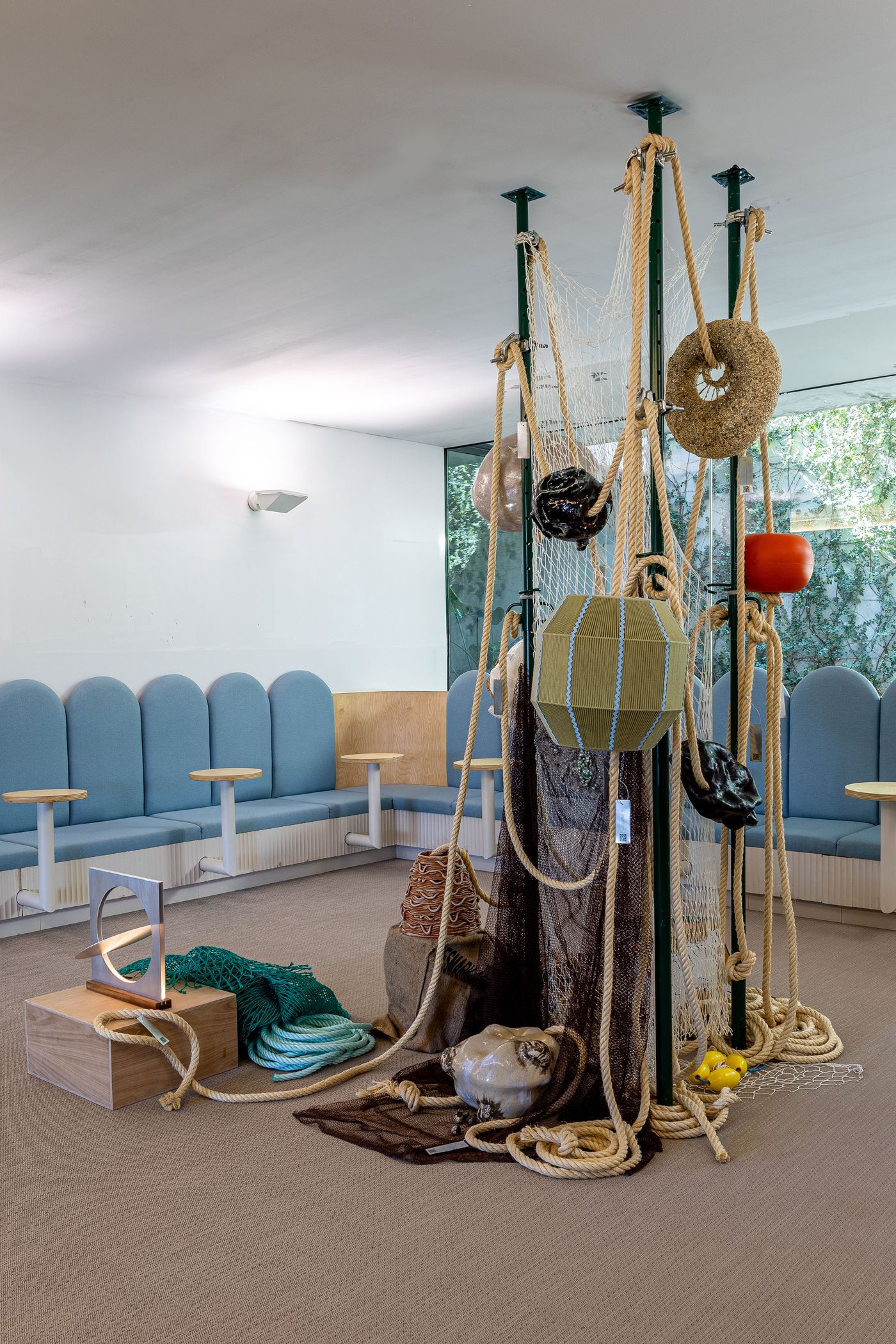 A view of LACS Santos' "The Tide Line Exhibition" captures an immersive, nature-inspired installation. Thick ropes, fishing nets, and marine elements like buoys cascade from central poles, grounded by a collection of objects including a bird-shaped sculpture. The textural, rustic art piece contrasts with the clean-lined, modern blue seating area and large windows revealing vibrant greenery.
