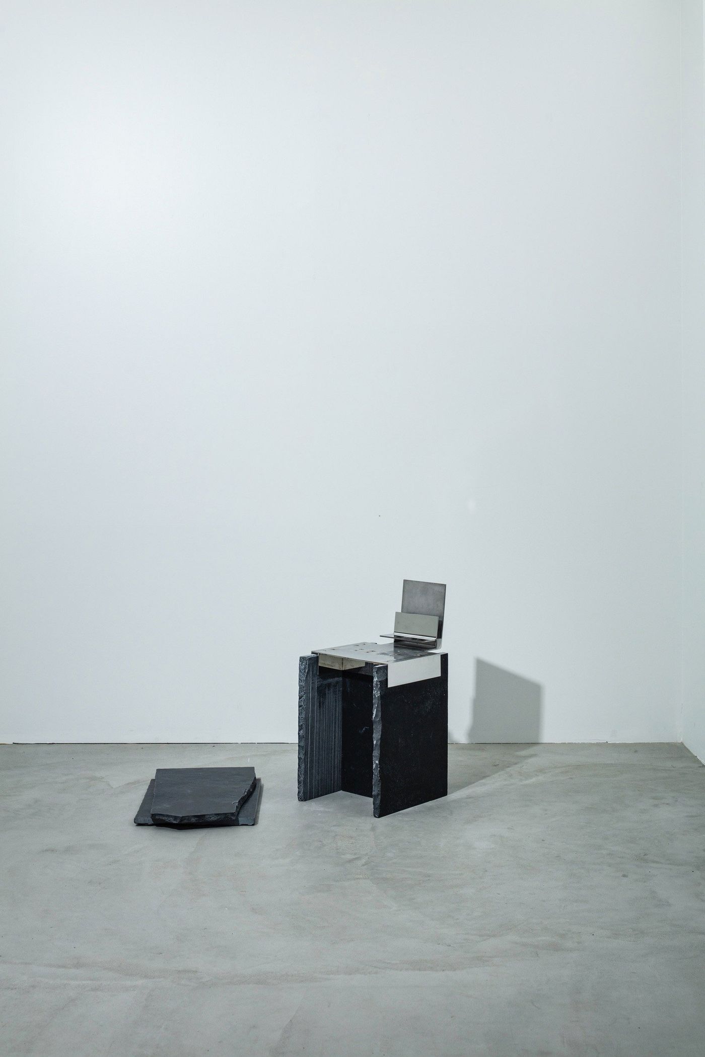 A minimalist, black stone chair with a metal seat, beside a flat stone slab, set against a plain white wall in an empty room.