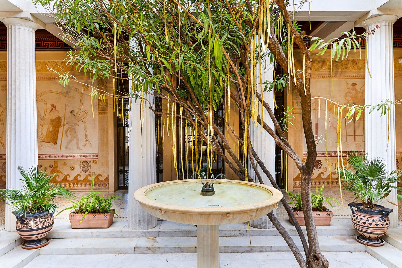 A wide view of the Villa Kerylos courtyard. A small water fountain sits in the middle of a square stone platform, flanked by a tree with branches from which golden metallic ribbons hang. The space is surrounded by large, white marble columns and neo-vernacular frescoes, with potted plants adding to the verdant and serene atmosphere. A wide view of the Villa Kerylos courtyard. A small water fountain sits in the middle of a square stone platform, flanked by a tree with branches from which golden metallic ribbons hang. The space is surrounded by large, white marble columns and neo-vernacular frescoes, with potted plants adding to the verdant and serene atmosphere.