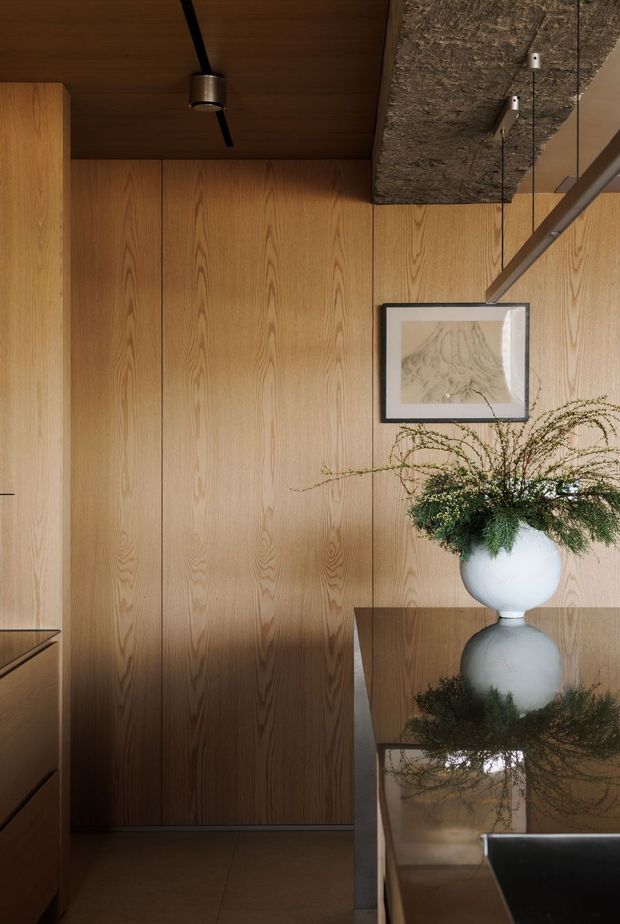 A study in refined textures and natural materials within a modern interior. The wall is clad in warm-toned wood paneling, contrasting with a raw concrete ceiling detail featuring exposed aggregate. A sleek, reflective surface on the counter holds a large white vase with verdant foliage, while a framed abstract artwork adds a subtle artistic touch. A study in refined textures and natural materials within a modern interior. The wall is clad in warm-toned wood paneling, contrasting with a raw concrete ceiling detail featuring exposed aggregate. A sleek, reflective surface on the counter holds a large white vase with verdant foliage, while a framed abstract artwork adds a subtle artistic touch.