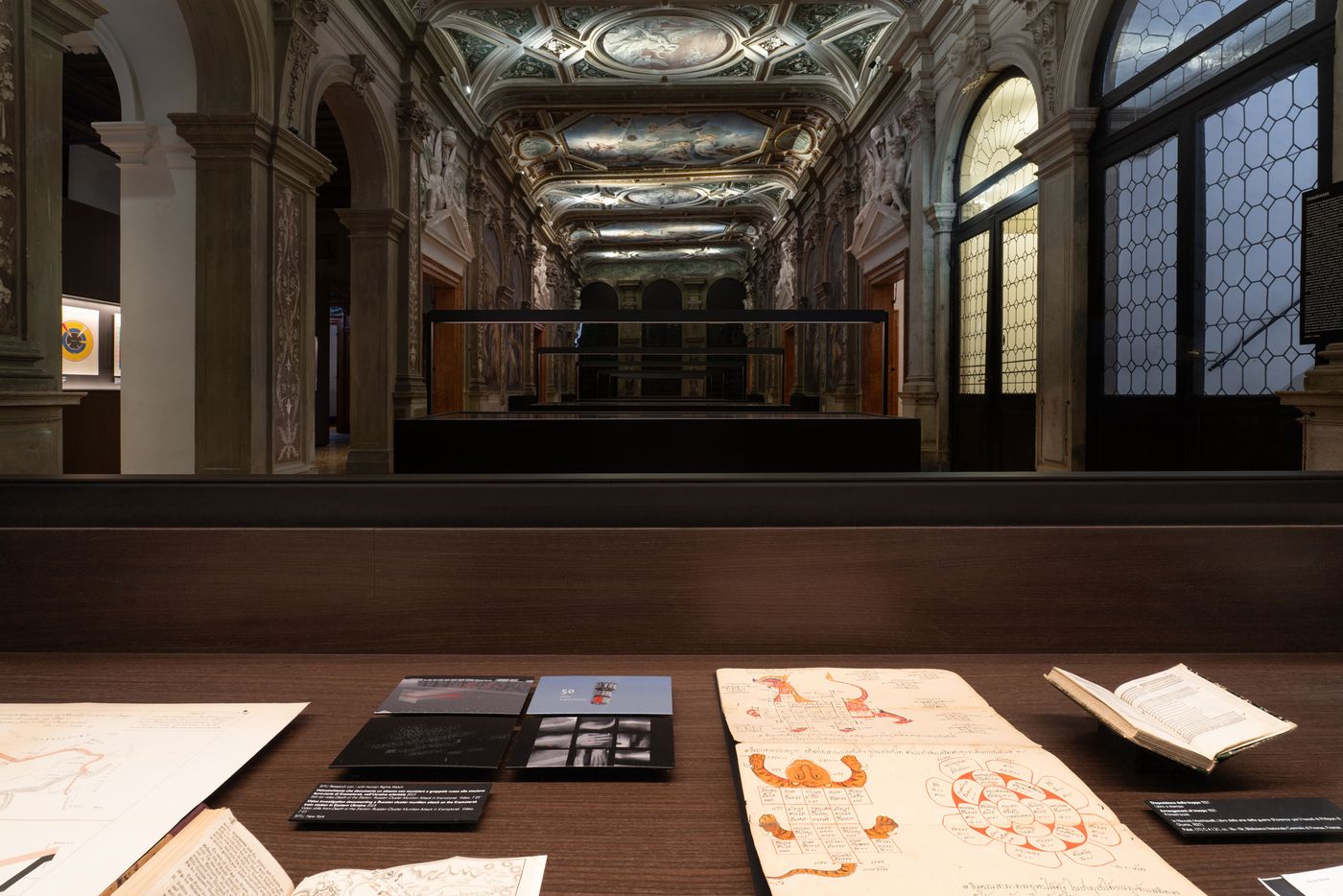 A richly decorated museum interior viewed from a wooden table displaying ancient manuscripts and notes, with ornate ceilings in the background.