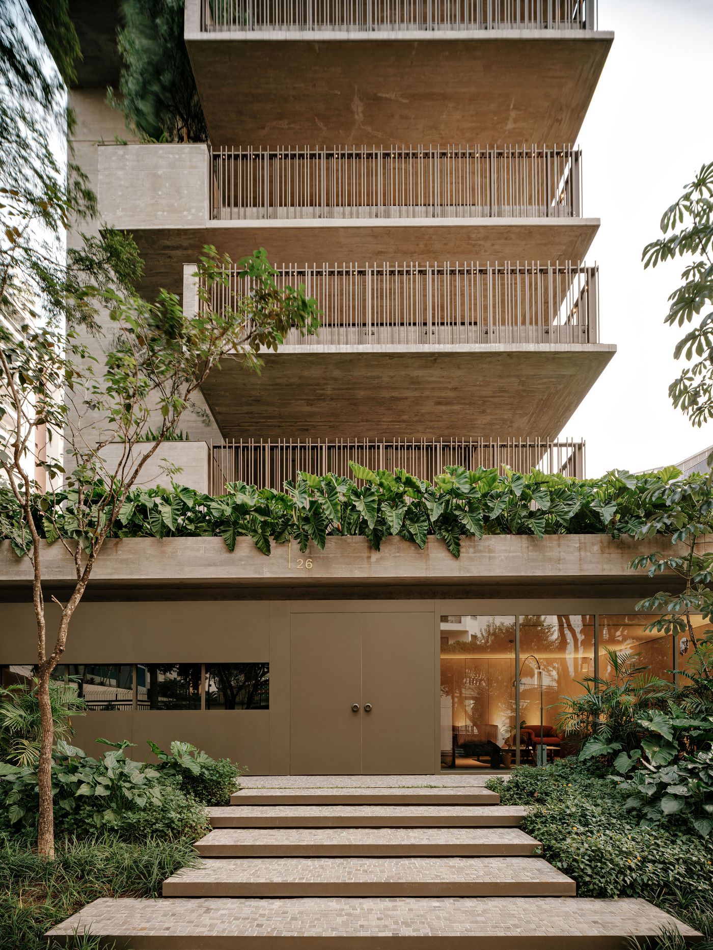 A welcoming entrance to the building, with a series of wide, shallow concrete steps leading to a recessed entryway. The raw concrete structure is softened by an abundance of diverse green plants, creating a sense of arrival that balances urban resilience with natural serenity, consistent with Studio Arthur Casas's design. A welcoming entrance to the building, with a series of wide, shallow concrete steps leading to a recessed entryway. The raw concrete structure is softened by an abundance of diverse green plants, creating a sense of arrival that balances urban resilience with natural serenity, consistent with Studio Arthur Casas's design.