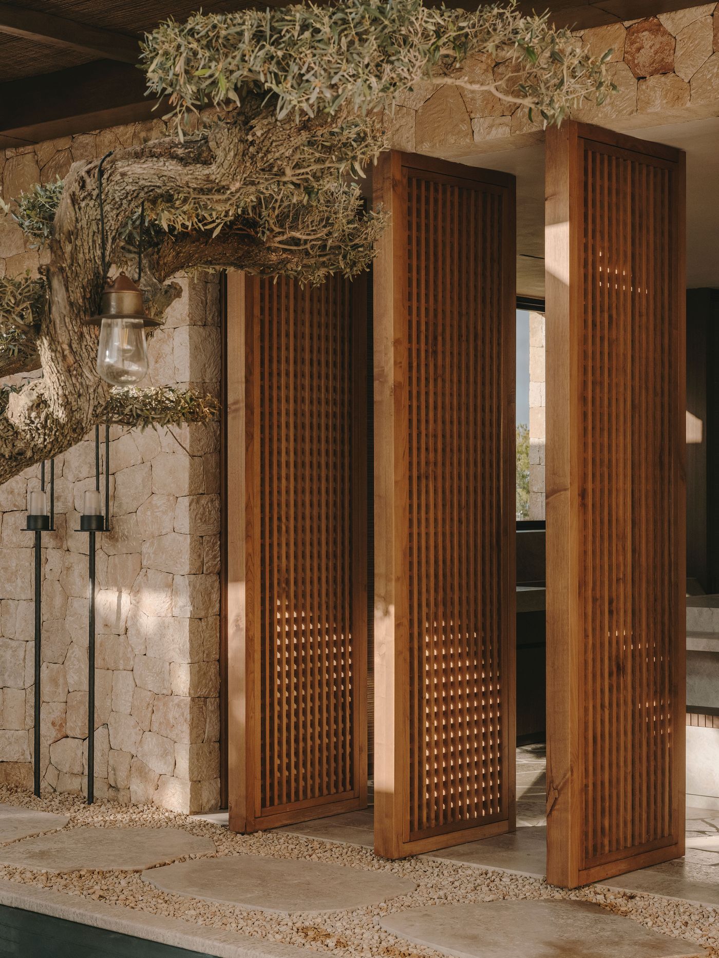 Wooden slatted doors stand beside a stone wall, with an olive tree and hanging lantern, creating a serene outdoor space.