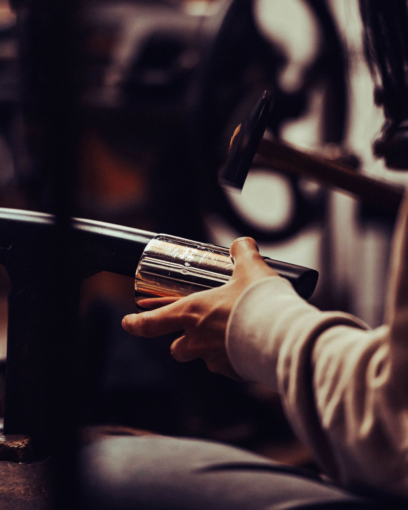 A close-up of a person shaping metal with a hammer, focusing on their hand holding a polished metal object in a workshop setting.