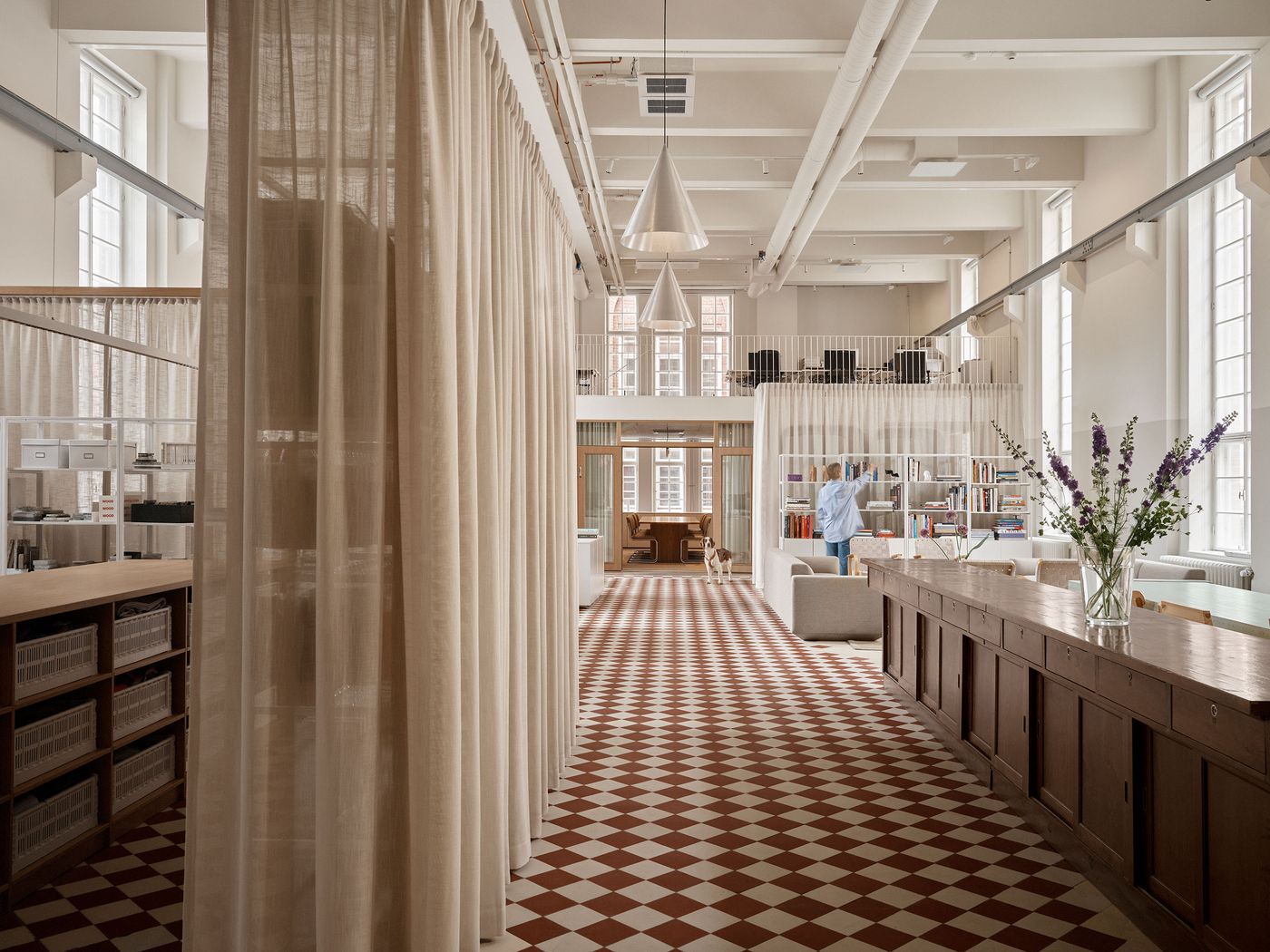 A long hallway with a distinctive red and white checkerboard floor leads through an open-plan office, defined by tall, flowing sheer curtains that create soft partitions. A person and a dog are visible near a minimalist white bookcase, hinting at a dynamic and inviting workspace. A long hallway with a distinctive red and white checkerboard floor leads through an open-plan office, defined by tall, flowing sheer curtains that create soft partitions. A person and a dog are visible near a minimalist white bookcase, hinting at a dynamic and inviting workspace.