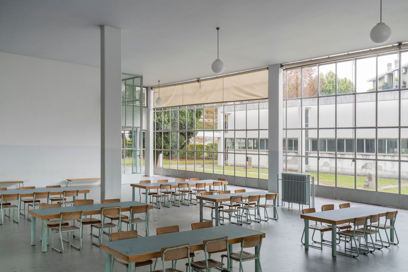 A large, open room with a high ceiling is furnished with rows of midcentury-modern school-style tables and chairs. The space is filled with a soft, diffused light from the expansive floor-to-ceiling windows and large globe pendant lights. The minimalist palette of white walls, light gray floor, and natural wood furniture creates a calm, functional atmosphere. The design is simple and industrial, reminiscent of a school cafeteria or a large shared workspace.