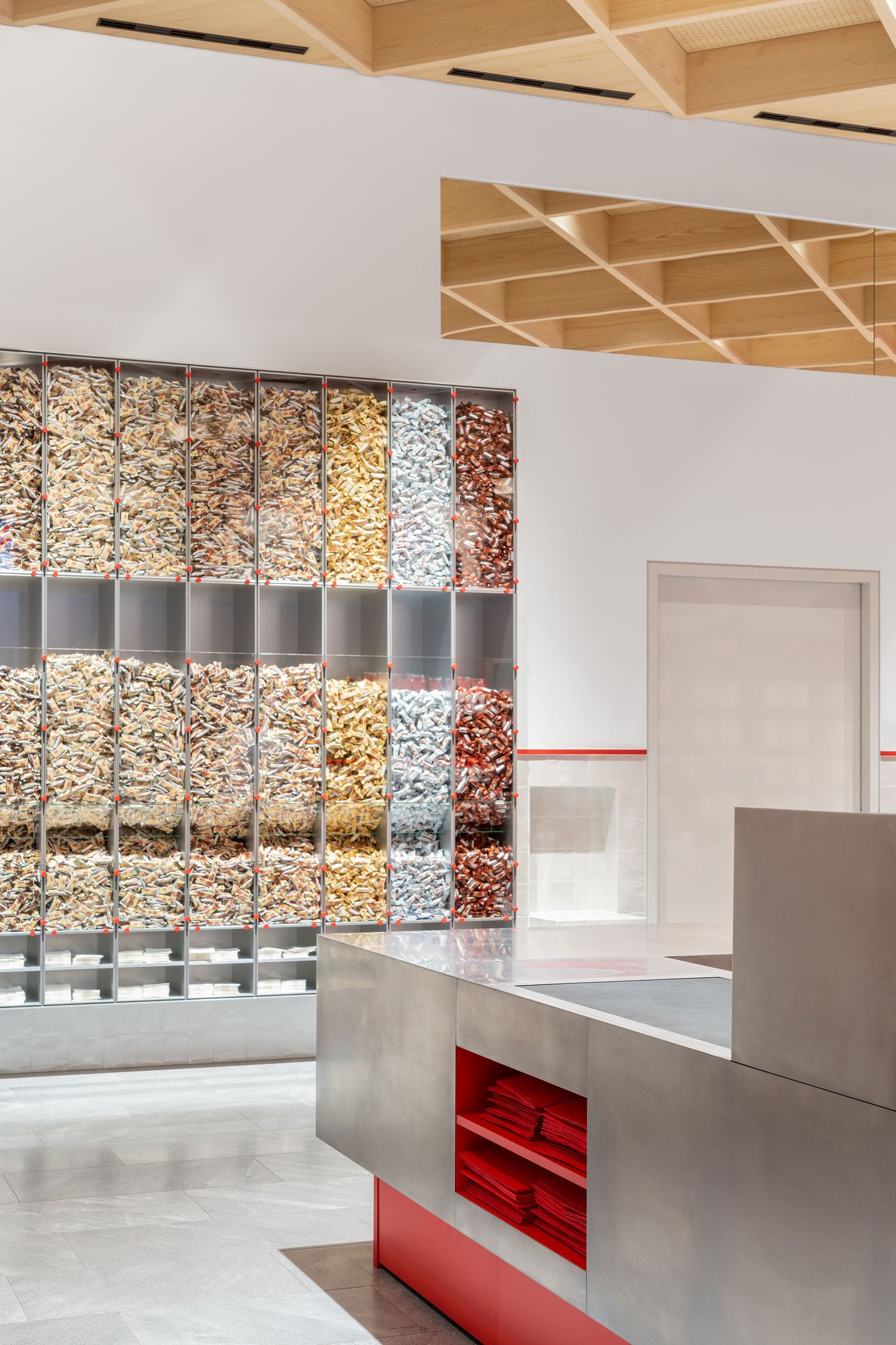 Floor-to-ceiling glazed display units reveal colorful product contents like an edible art installation. The systematic grid organization transforms retail storage into architectural spectacle, while warm timber ceiling elements soften the clinical precision of the transparent merchandising system below Floor-to-ceiling glazed display units reveal colorful product contents like an edible art installation. The systematic grid organization transforms retail storage into architectural spectacle, while warm timber ceiling elements soften the clinical precision of the transparent merchandising system below