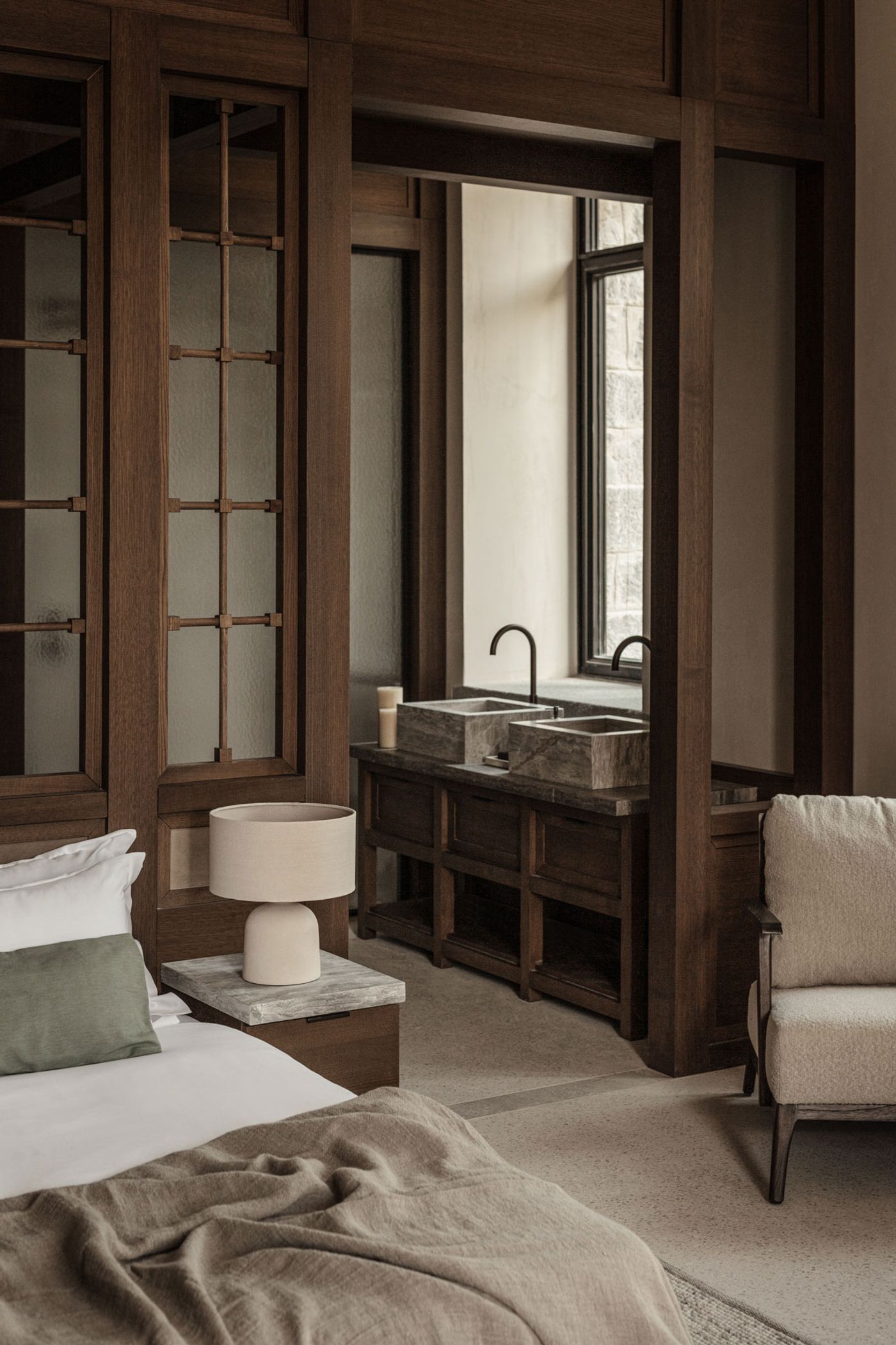 A sophisticated Japandi-inspired bedroom transitions into a bathroom area, defined by rich, dark timber-framed partitions with subtle grid patterns. Twin stone vessel sinks with elegant black faucets stand on a dark wood vanity, illuminated by natural light from a large window, creating a cohesive, warm, and inviting atmosphere. A sophisticated Japandi-inspired bedroom transitions into a bathroom area, defined by rich, dark timber-framed partitions with subtle grid patterns. Twin stone vessel sinks with elegant black faucets stand on a dark wood vanity, illuminated by natural light from a large window, creating a cohesive, warm, and inviting atmosphere.