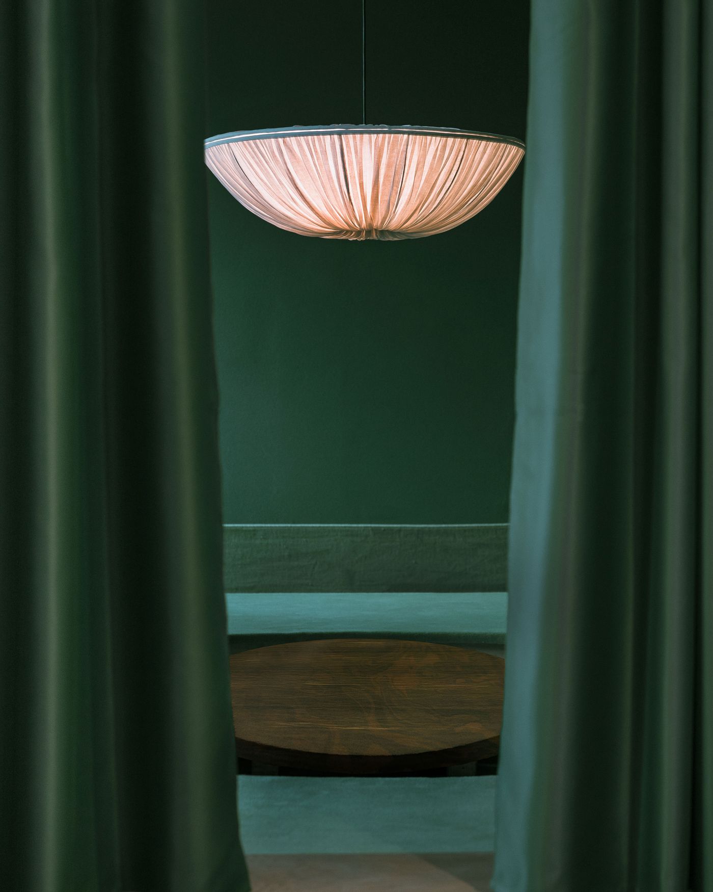 A softly glowing chandelier hangs between two green curtains, illuminating a circular wooden table against a deep green backdrop.