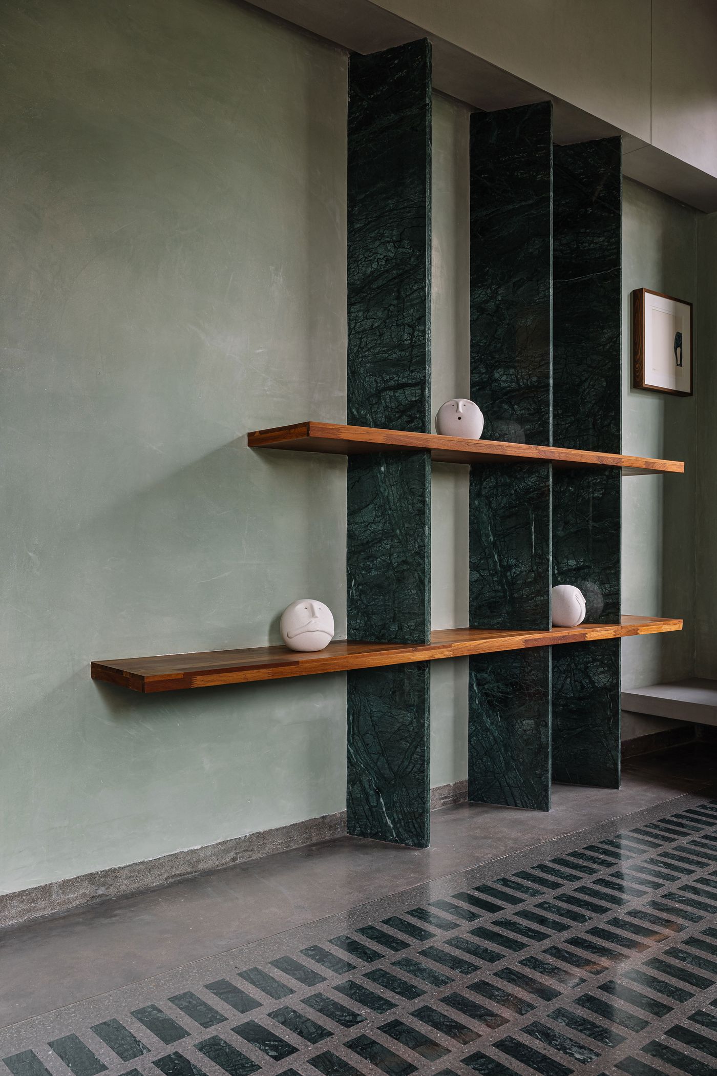 A modern shelving unit, featuring thick wooden shelves supported by tall, dark green marble slabs. Three white spherical decorative objects are symmetrically placed on the shelves. The wall behind is a muted green, while the floor is a grey tile with a dark green geometric pattern, creating an elegant blend of natural materials and contemporary design.