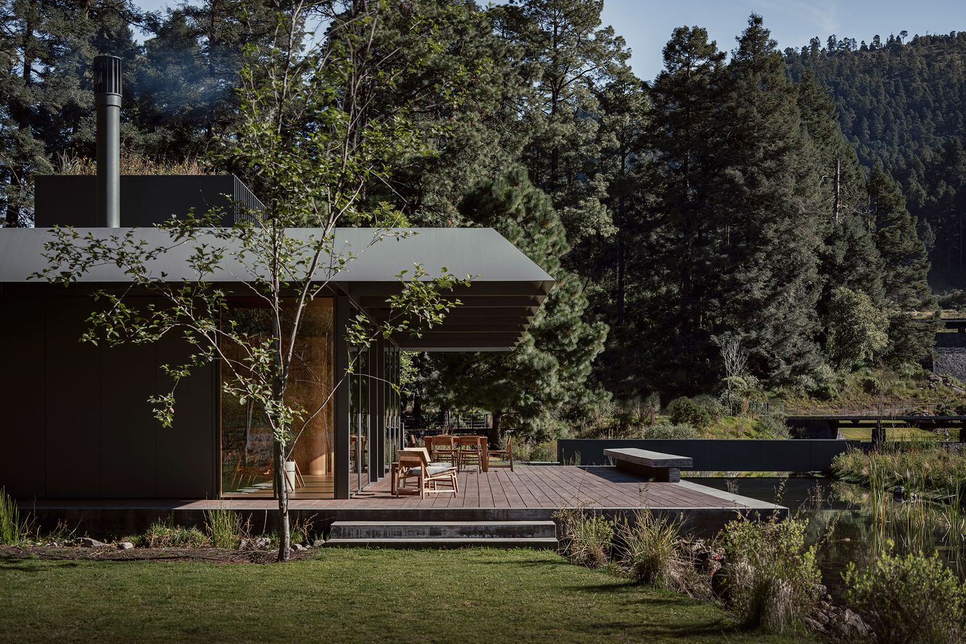 A modern cabin nestled among trees, featuring a wooden deck, outdoor seating, and a tranquil pond, creating a serene nature retreat.