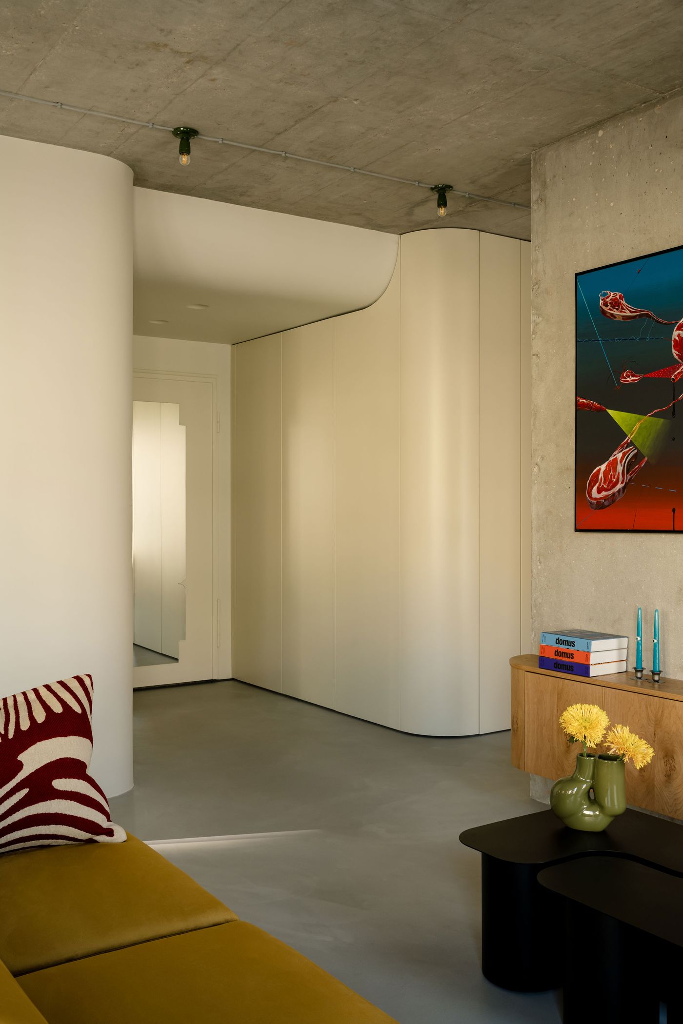 This transitional view captures the minimalist sophistication of the hallway, where curving cabinetry and creamy walls offset the industrial ceiling. A sculptural mirror and zebra-striped cushion add playful punctuation to the otherwise serene, streamlined composition.
