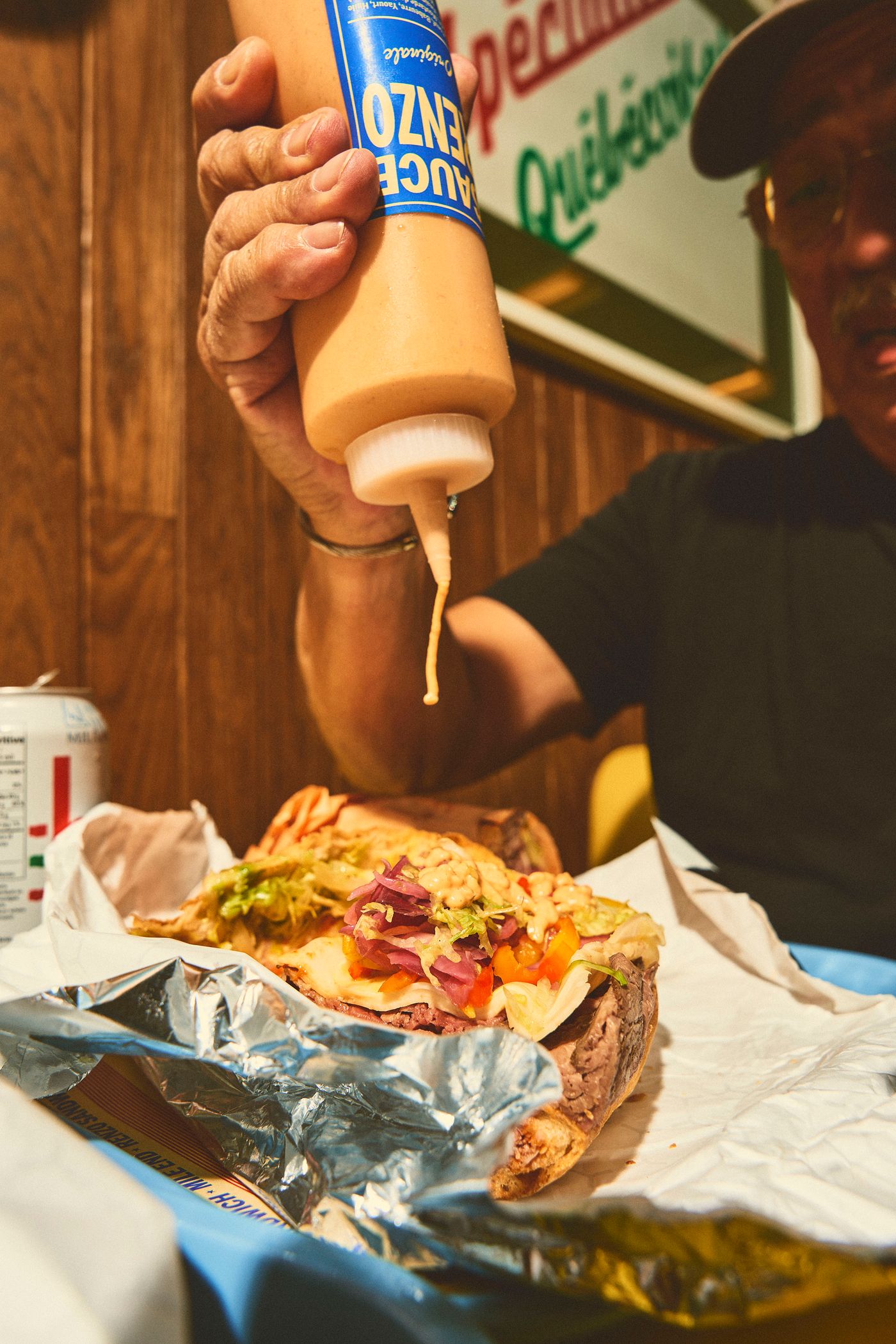 A hand holds a bottle of "SAUCE ORIGINALE RENZO" over a large sandwich, ready to add a topping. The sandwich, wrapped in foil, is piled high with meats and vegetables. The scene captures a moment of preparation and indulgence, characteristic of a classic sandwich shop experience.