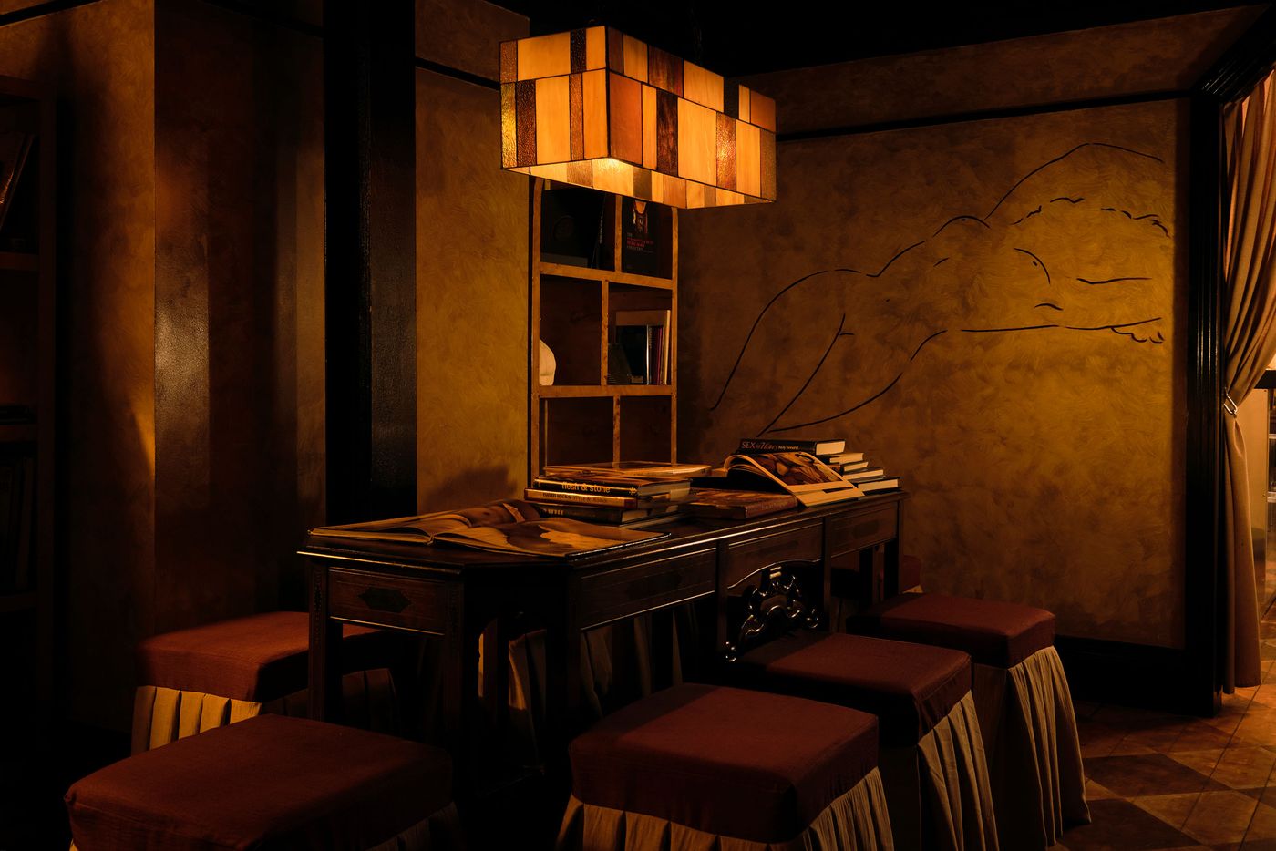 A dramatic, eclectic corner within The Roses of Elagabalus features a prominent wooden box-like pendant light casting a warm glow over a dark, ornate wooden desk piled with books. Four plush, skirted stools in deep red are arranged around it, set against a gold-toned wall with an abstract outline, creating a luxurious and intellectual atmosphere. A dramatic, eclectic corner within The Roses of Elagabalus features a prominent wooden box-like pendant light casting a warm glow over a dark, ornate wooden desk piled with books. Four plush, skirted stools in deep red are arranged around it, set against a gold-toned wall with an abstract outline, creating a luxurious and intellectual atmosphere.