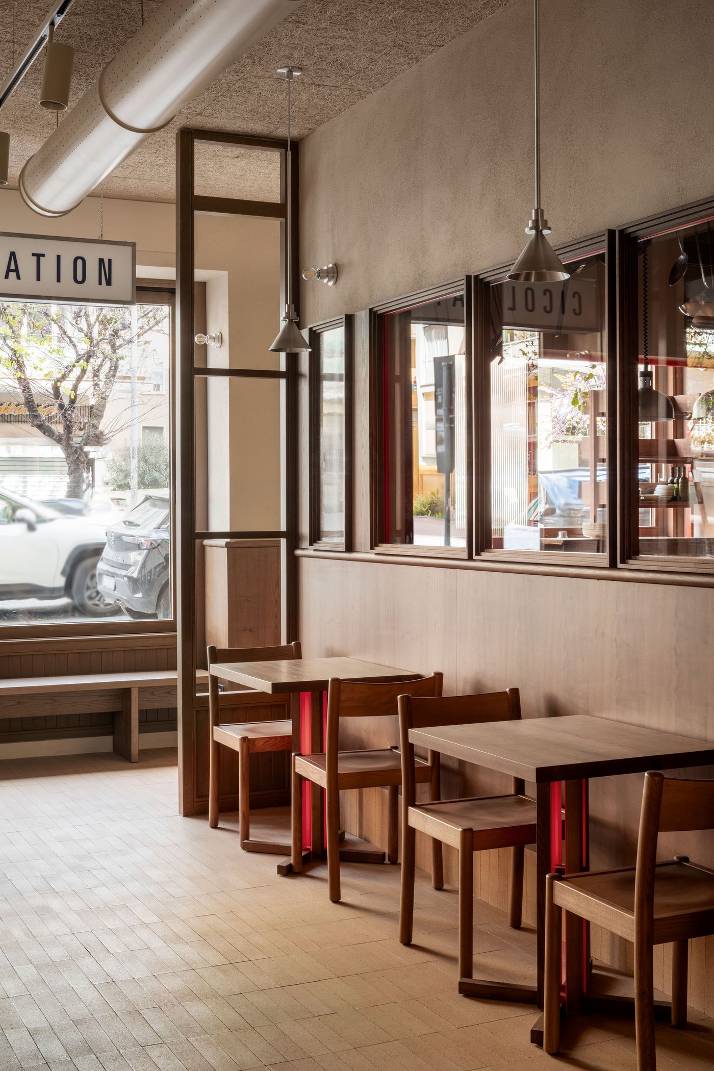 A cozy, well-lit corner of Santo Palato by Naessi Studio, featuring intimate dining nooks. Each wooden table with a subtle red base is paired with simple wooden chairs. The space is anchored by warm wood wainscoting and framed windows offering glimpses of the outside, creating a relaxed, authentic Roman trattoria feel.