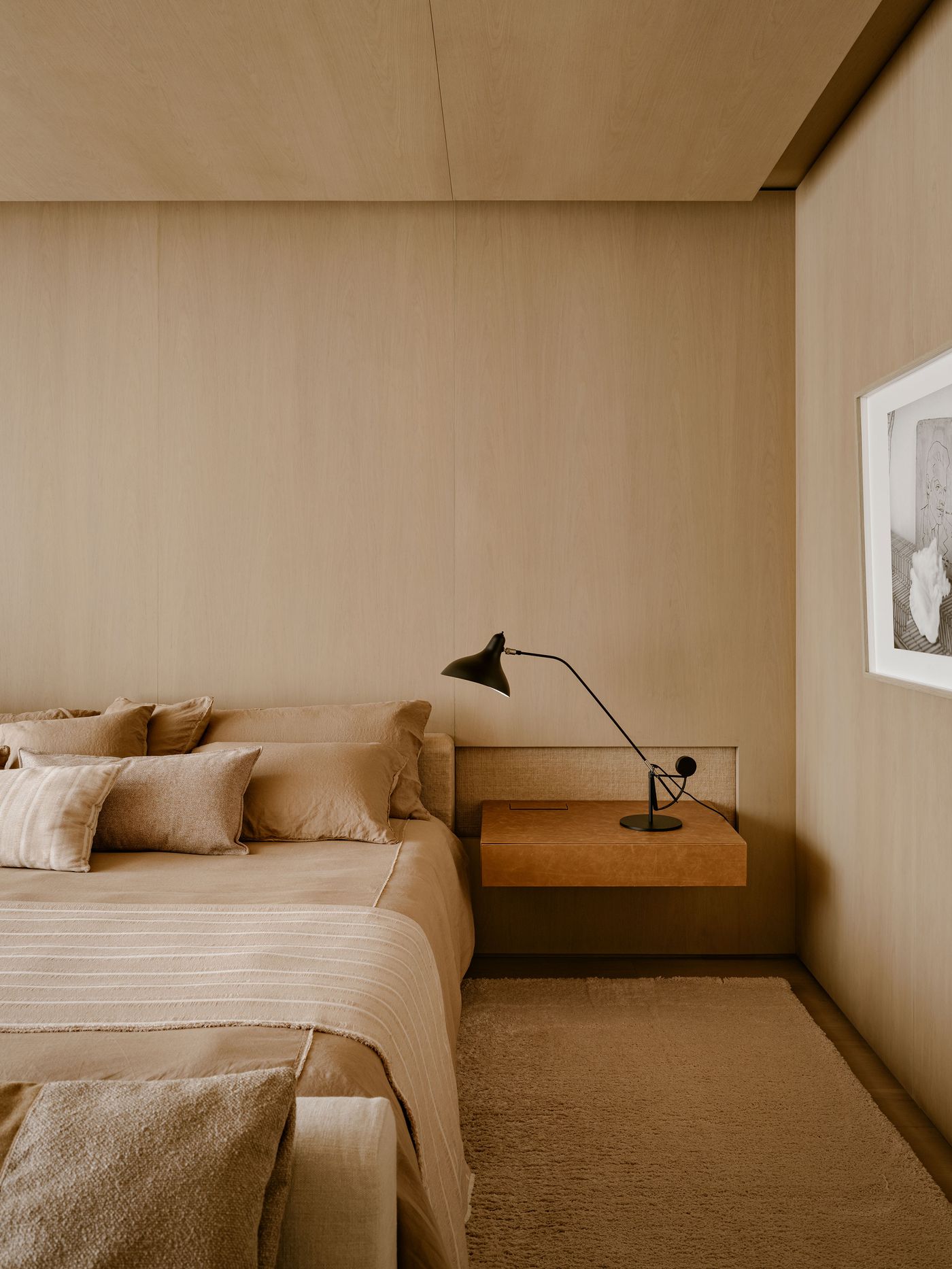 Modern bedroom featuring a beige color palette, plush bedding, a minimal lamp, and a wooden nightstand against a wood-paneled wall.