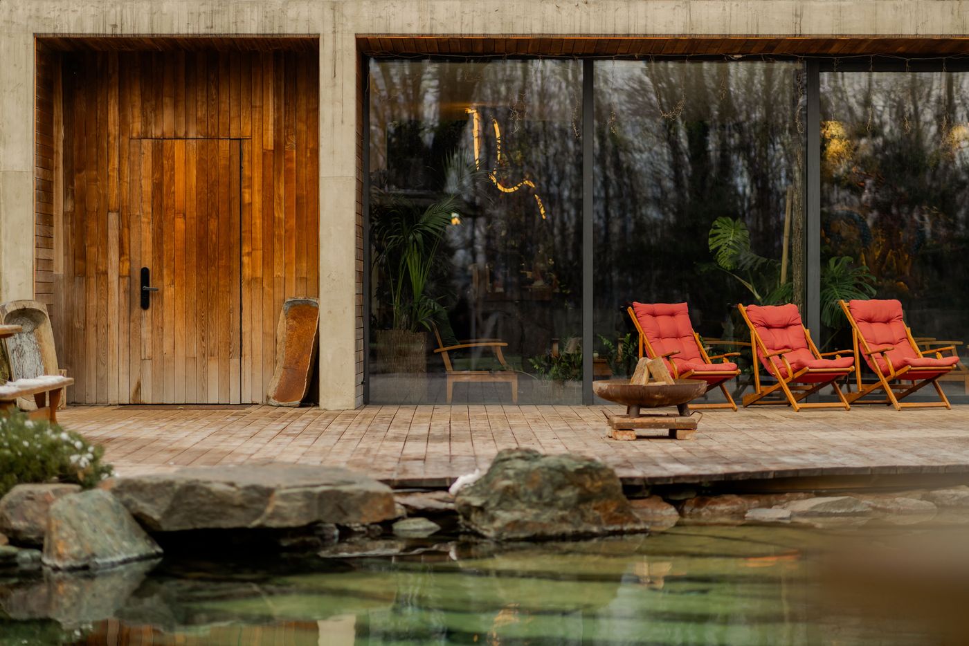 Modern wooden cabin featuring a glass wall, three red lounge chairs on a wooden deck, and a serene pond with rocks. Modern wooden cabin featuring a glass wall, three red lounge chairs on a wooden deck, and a serene pond with rocks.