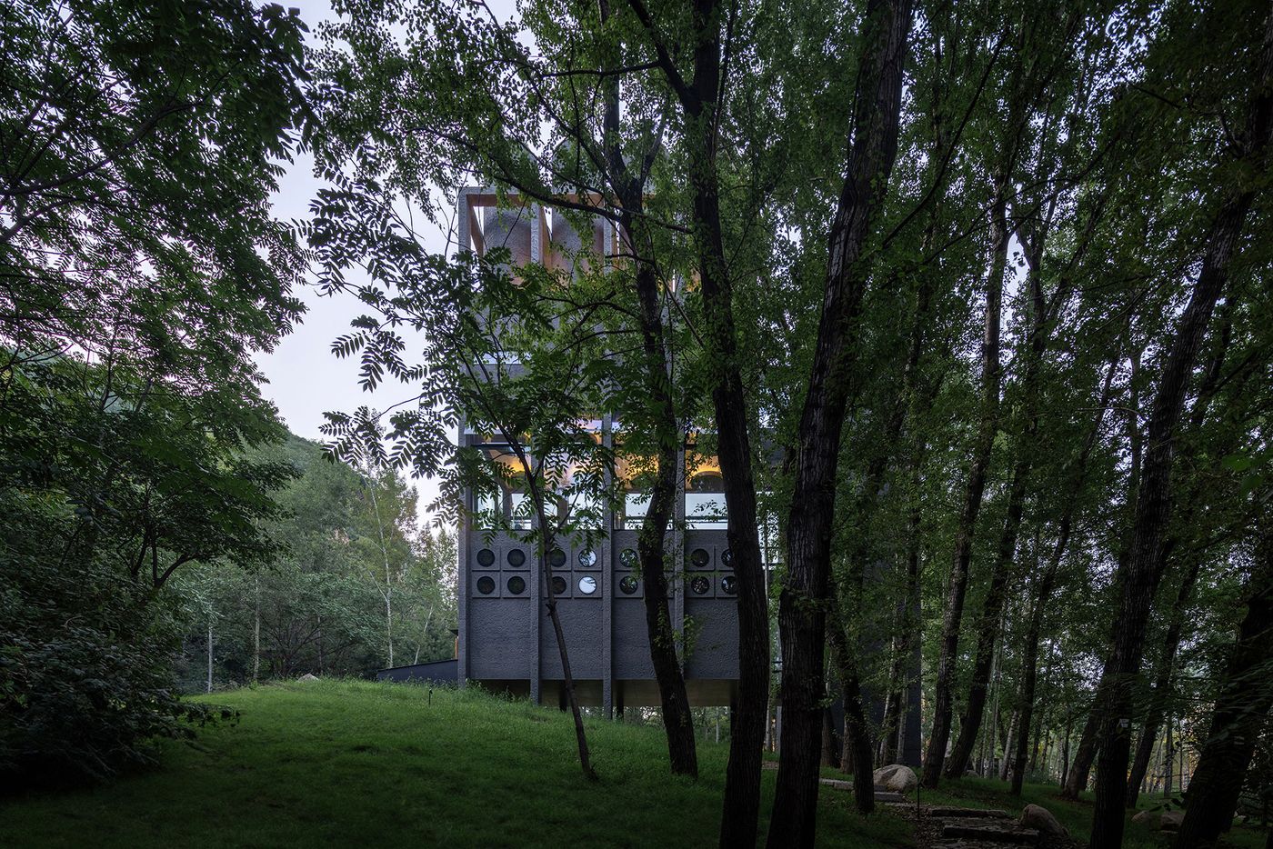 A modern, multi-level building partially obscured by lush green trees in a serene forest setting, illuminated by warm interior lights.