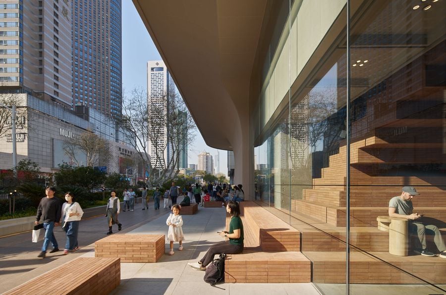 A bustling urban scene with people walking along a sidewalk, modern buildings, and a glass storefront featuring wooden benches.