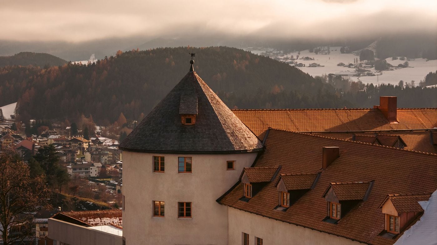 A round tower with a conical wooden-shingle roof and a weathervane rises above Castel Badia's terracotta-tiled roofs and timber dormers. Late-afternoon light warms the rendered walls, while larch-covered hills, snow-dusted fields and the village below recede into low cloud across the Puster Valley. A round tower with a conical wooden-shingle roof and a weathervane rises above Castel Badia's terracotta-tiled roofs and timber dormers. Late-afternoon light warms the rendered walls, while larch-covered hills, snow-dusted fields and the village below recede into low cloud across the Puster Valley.