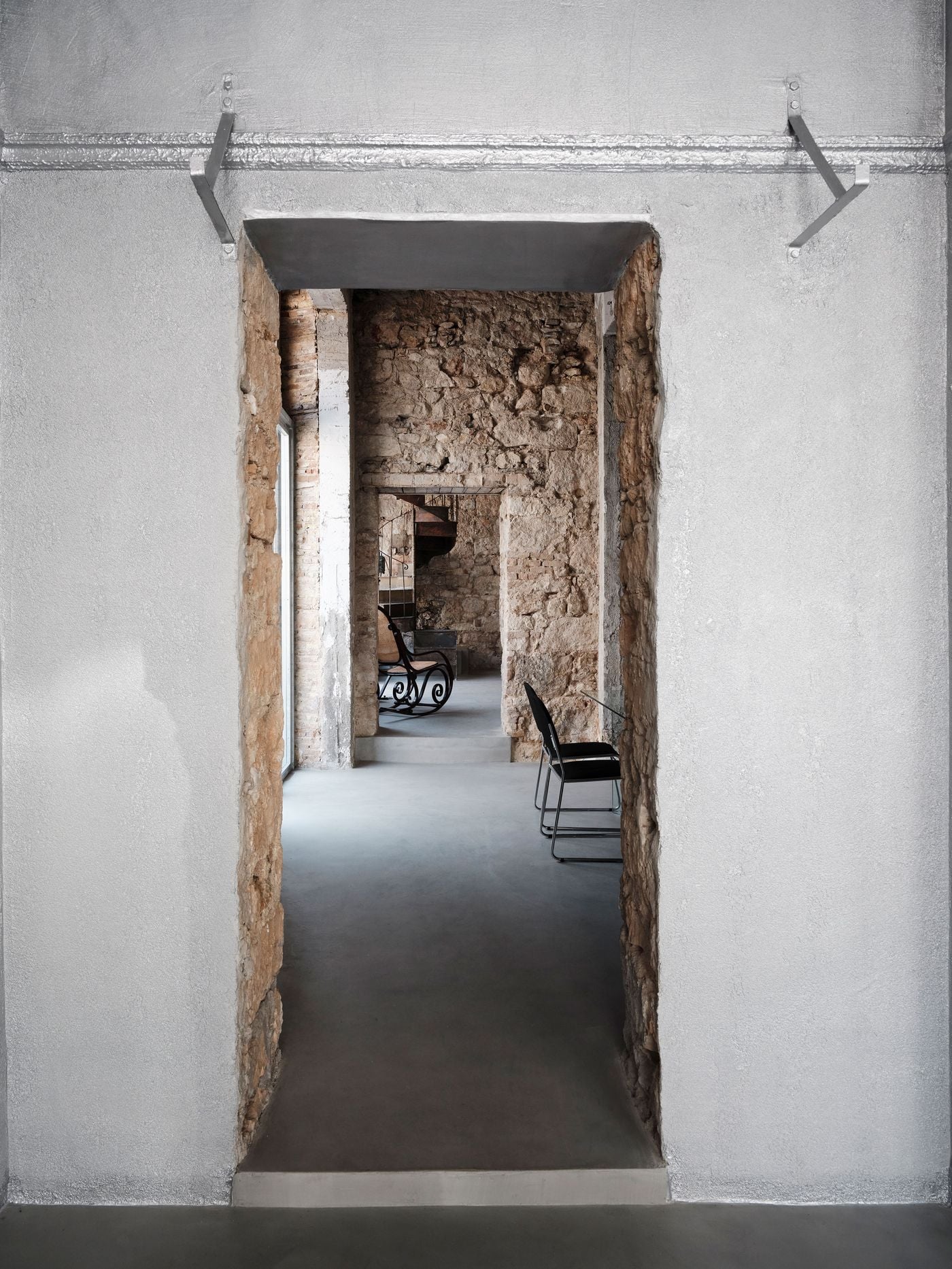 A silvery, liquid-metal-finished wall frames a rough-edged opening onto the building’s older masonry interior. The long axial view layers smooth contemporary surfaces with exposed stone, while glimpses of black chairs and a cane rocking chair draw the eye through a succession of interconnected spaces. A silvery, liquid-metal-finished wall frames a rough-edged opening onto the building’s older masonry interior. The long axial view layers smooth contemporary surfaces with exposed stone, while glimpses of black chairs and a cane rocking chair draw the eye through a succession of interconnected spaces.