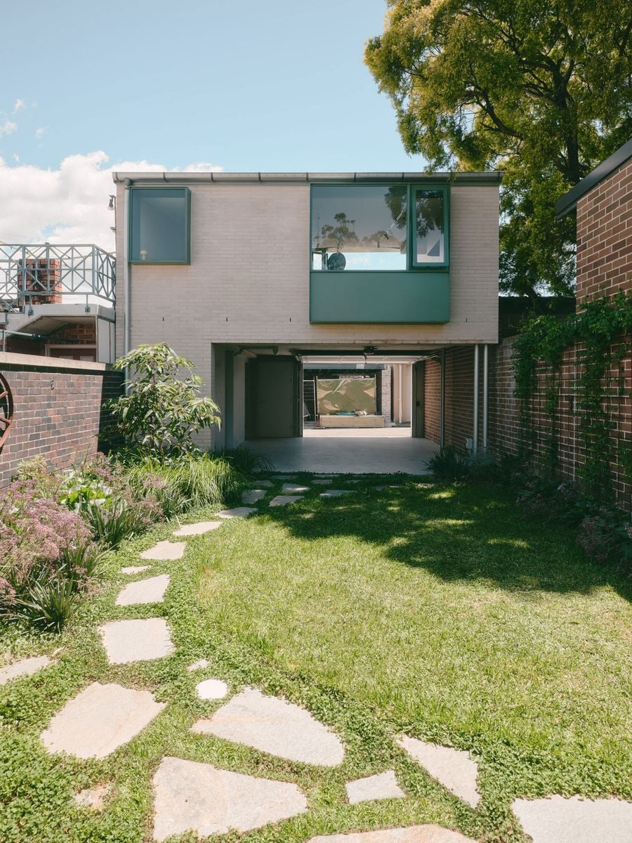 Seen from the garden, the addition takes on a quieter character, with pale brickwork and a broad sage-green framed window overlooking a grassy backyard. The ground-floor garage opens completely beneath the studio, creating a sheltered passage and flexible social space that visually connects garden and laneway. Seen from the garden, the addition takes on a quieter character, with pale brickwork and a broad sage-green framed window overlooking a grassy backyard. The ground-floor garage opens completely beneath the studio, creating a sheltered passage and flexible social space that visually connects garden and laneway.