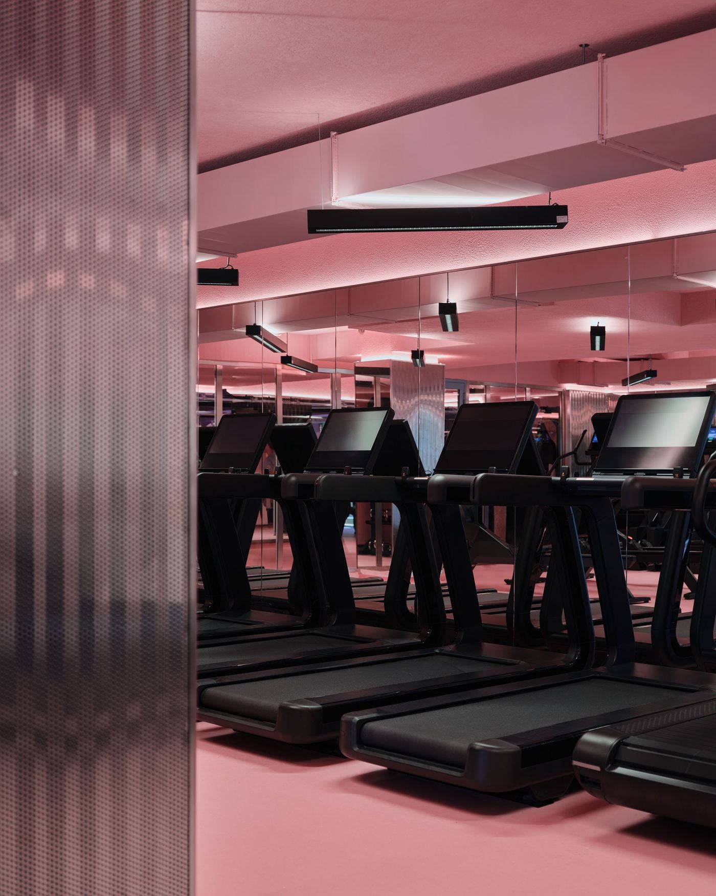 Rows of black treadmills line the Ken Club’s cardio area, set against a pink floor and ceiling glow that softens the otherwise utilitarian equipment. Mirrored walls multiply the space, while perforated metal screens and elongated black light fittings reinforce the club’s sleek, industrial-meets-playful material language. Rows of black treadmills line the Ken Club’s cardio area, set against a pink floor and ceiling glow that softens the otherwise utilitarian equipment. Mirrored walls multiply the space, while perforated metal screens and elongated black light fittings reinforce the club’s sleek, industrial-meets-playful material language.