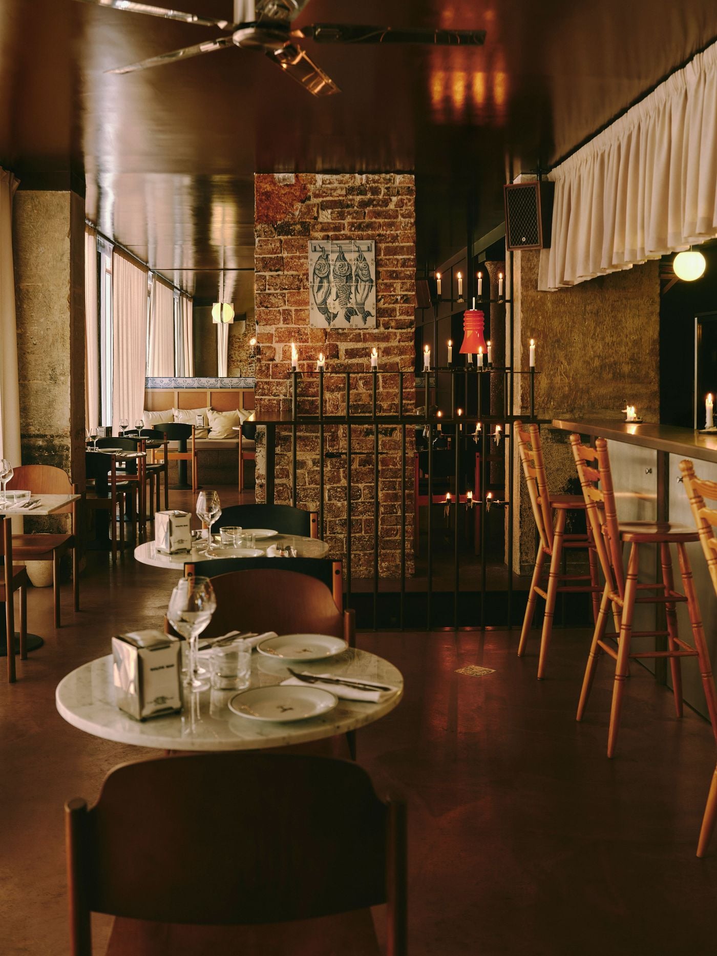 Round marble tables lead the eye through SaltoBar in Paris towards a brick pier hung with a tiled panel of three hanging fish. A black metal railing crowded with candles and a scarlet pendant separates the dining room from the bar. There, turned-pine stools line a steel counter beneath a pleated cream valance. Round marble tables lead the eye through SaltoBar in Paris towards a brick pier hung with a tiled panel of three hanging fish. A black metal railing crowded with candles and a scarlet pendant separates the dining room from the bar. There, turned-pine stools line a steel counter beneath a pleated cream valance.