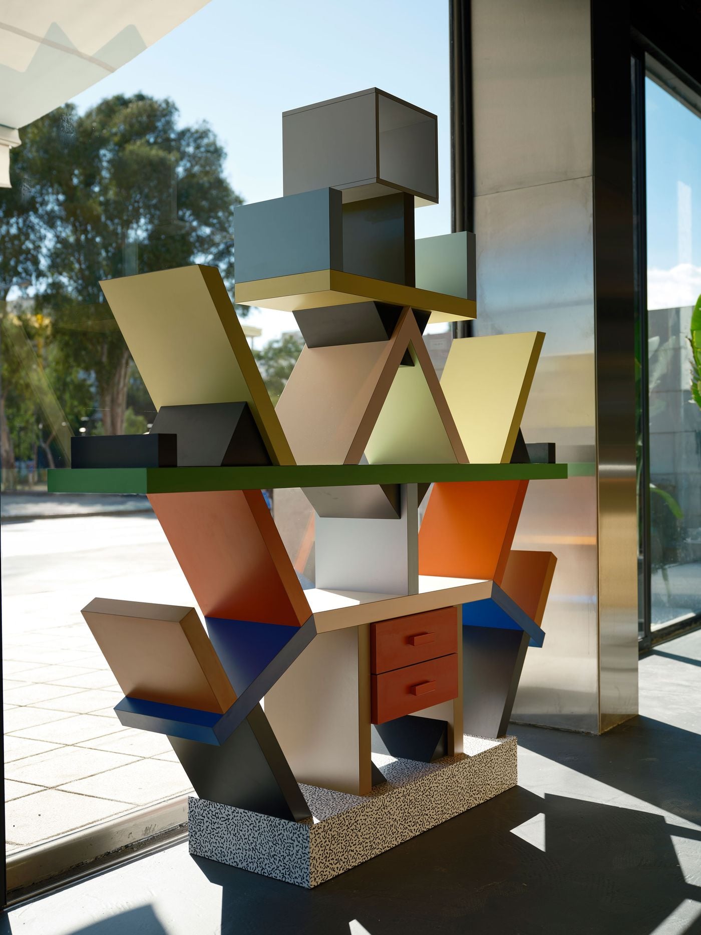 Ettore Sottsass's 1981 Carlton bookcase stands in sunlight by the window at Seth & Manni's. Its angled shelves in yellow, orange, blue and grey rise from a black-and-white patterned laminate base. The piece casts crisp geometric shadows across the floor beside a stainless-steel column Ettore Sottsass's 1981 Carlton bookcase stands in sunlight by the window at Seth & Manni's. Its angled shelves in yellow, orange, blue and grey rise from a black-and-white patterned laminate base. The piece casts crisp geometric shadows across the floor beside a stainless-steel column