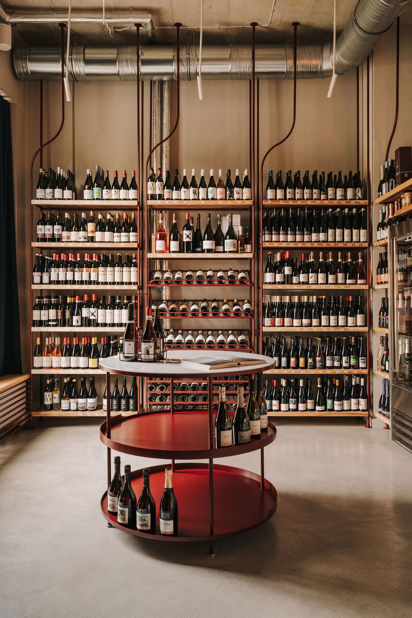 The ground-floor wine shop at WIN in Warsaw features extensive timber shelving filled with bottles, framed by slender burgundy metal supports that curve organically towards the ceiling. A circular, three-tiered display combines burgundy metalwork with a white tabletop, while exposed ventilation ducts, raw concrete and pale flooring reinforce the space’s refined industrial aesthetic. The ground-floor wine shop at WIN in Warsaw features extensive timber shelving filled with bottles, framed by slender burgundy metal supports that curve organically towards the ceiling. A circular, three-tiered display combines burgundy metalwork with a white tabletop, while exposed ventilation ducts, raw concrete and pale flooring reinforce the space’s refined industrial aesthetic.