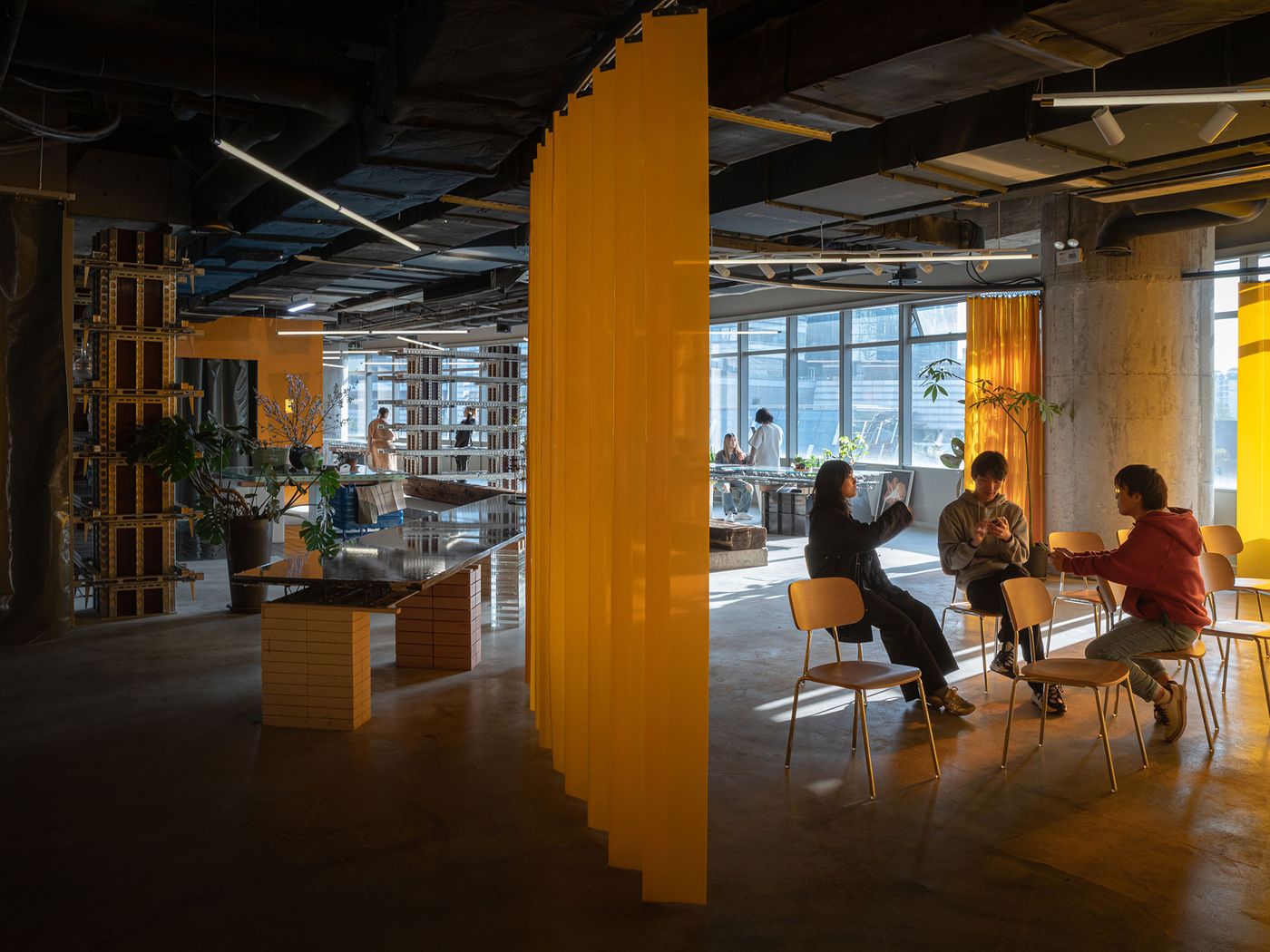 This image of 889 GLO Art Space reveals an open, industrial-style interior with a bold yellow curtain providing a striking vertical accent. Groups of people are engaged in casual interactions, seated on simple chairs, under abundant natural light streaming through large windows, enhancing the space's adaptable and inviting public art gallery ambiance.