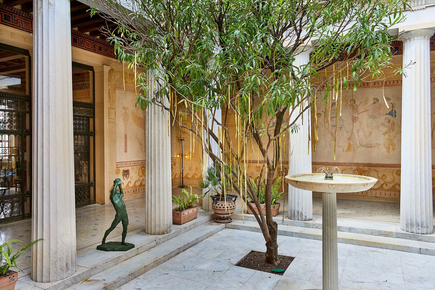 A sunlit corner of the Villa Kerylos courtyard, where a small bronze statue of a figure stands in front of a classical column. In the background, a tree dangles golden, ribbon-like metallic strips over a central fountain. The space blends biophilic elements with traditional Greek and Roman architectural features. A sunlit corner of the Villa Kerylos courtyard, where a small bronze statue of a figure stands in front of a classical column. In the background, a tree dangles golden, ribbon-like metallic strips over a central fountain. The space blends biophilic elements with traditional Greek and Roman architectural features.