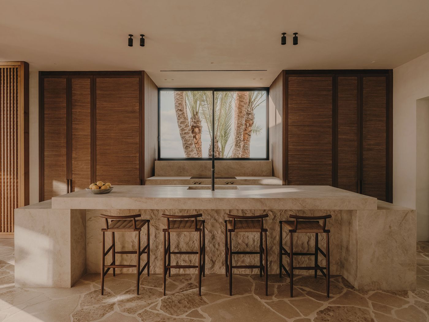 Modern kitchen with a stone island, wooden cabinets, bar stools, and a large window showcasing palm trees outside. Modern kitchen with a stone island, wooden cabinets, bar stools, and a large window showcasing palm trees outside.