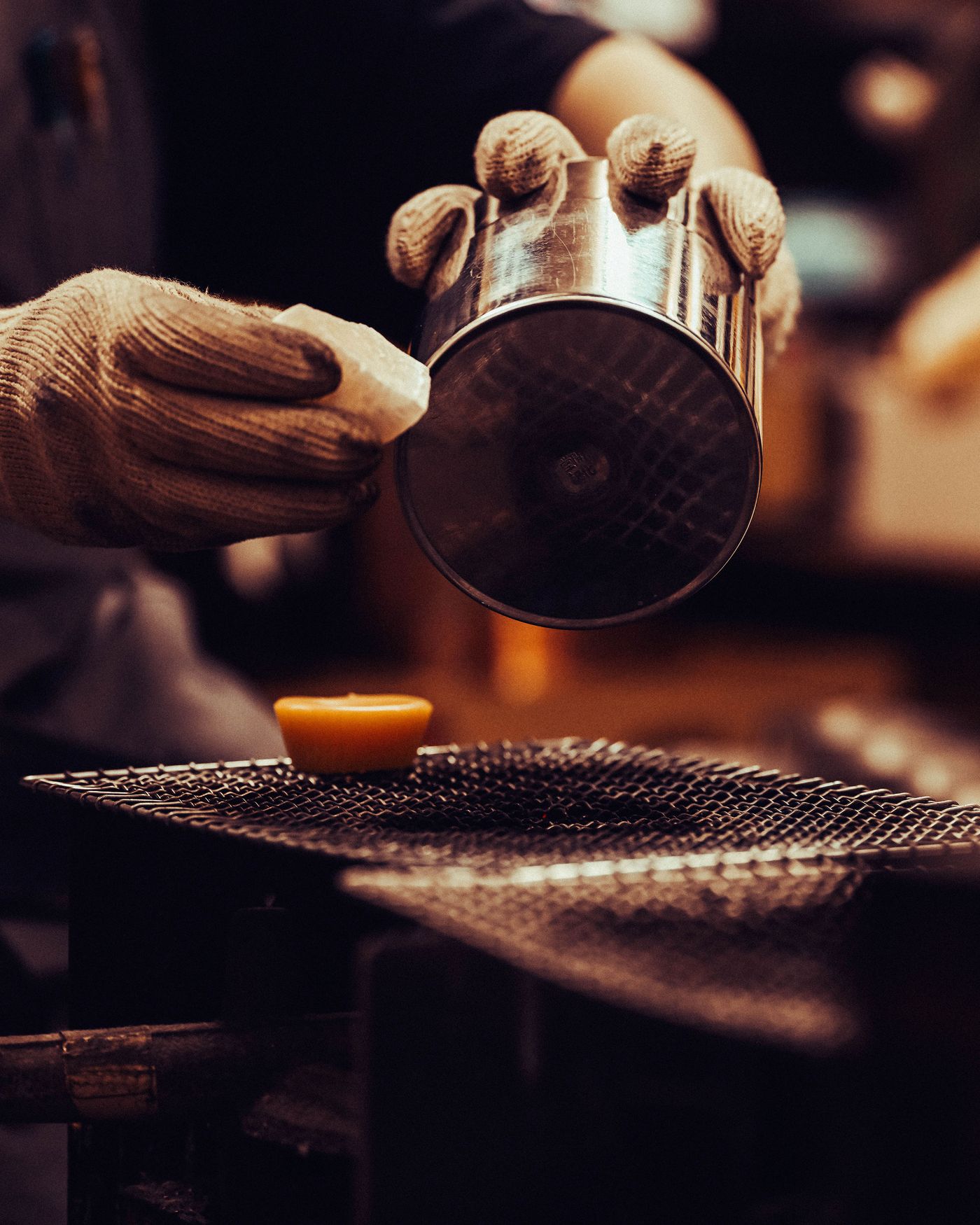 A gloved hand lifts a shiny metal container above an orange round object resting on a mesh surface in a workshop setting. A gloved hand lifts a shiny metal container above an orange round object resting on a mesh surface in a workshop setting.