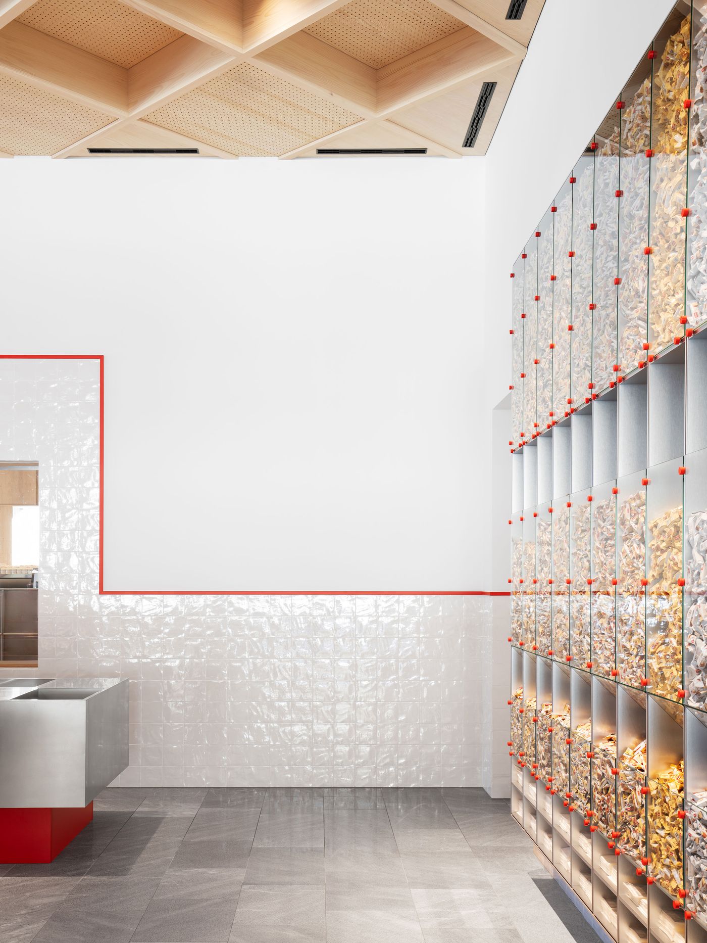 Cascading glass vessels create a kinetic wall installation, transforming product display into dynamic sculpture. Red accent dots punctuate the transparent grid system, while the geometric ceiling treatment above provides architectural counterpoint to the organic flow of contents within each vessel.