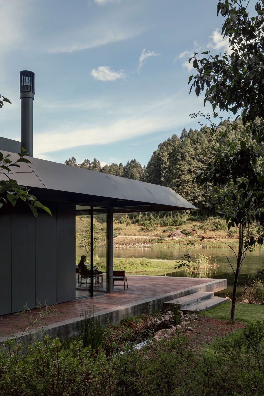 A modern lakeside cabin with a large overhanging roof, surrounded by greenery, features a person sitting on a deck, reading peacefully.