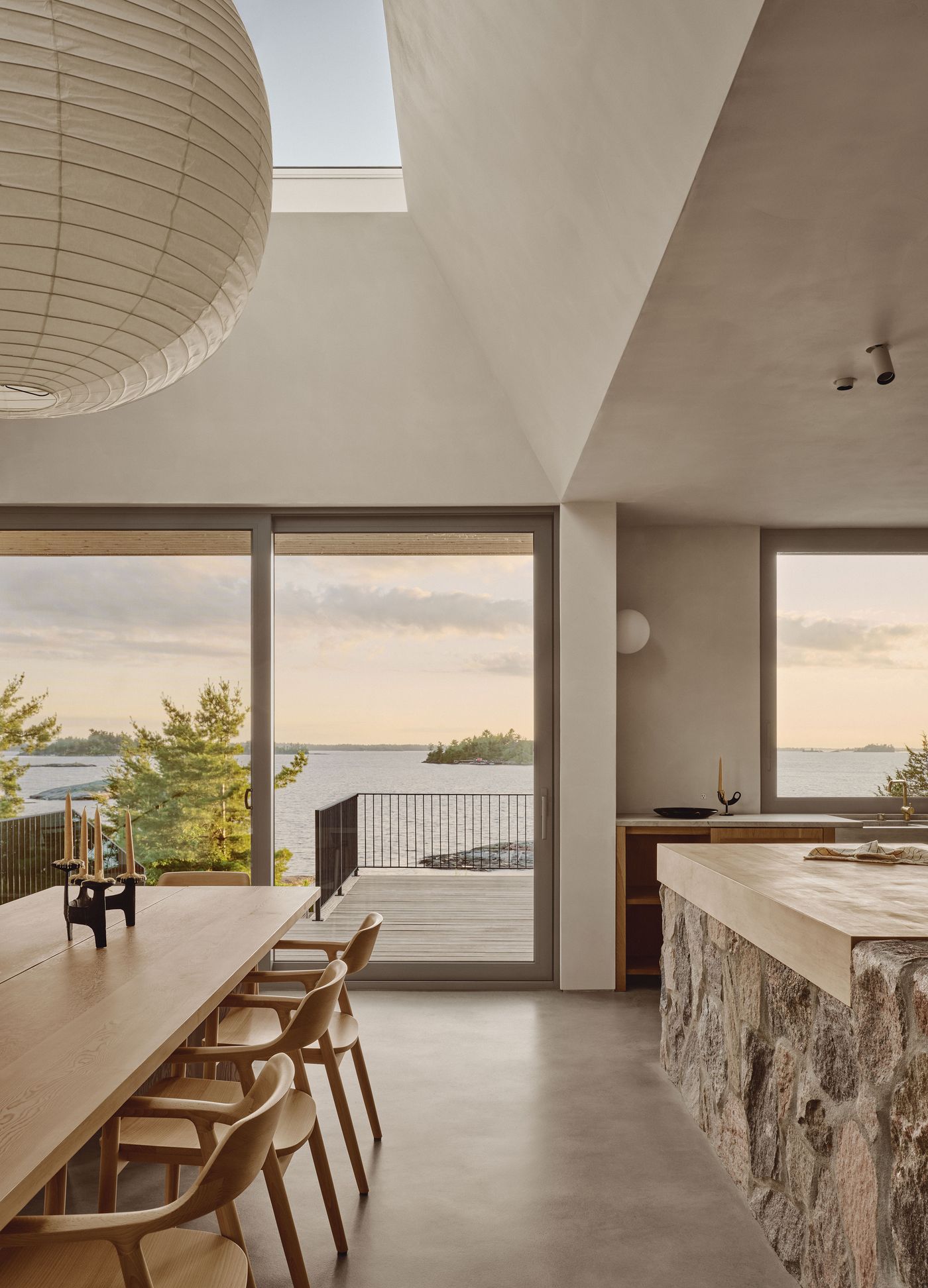 Modern kitchen-dining area with a stone island, wooden table, and large windows offering a serene view of the lake and sunset. Modern kitchen-dining area with a stone island, wooden table, and large windows offering a serene view of the lake and sunset.