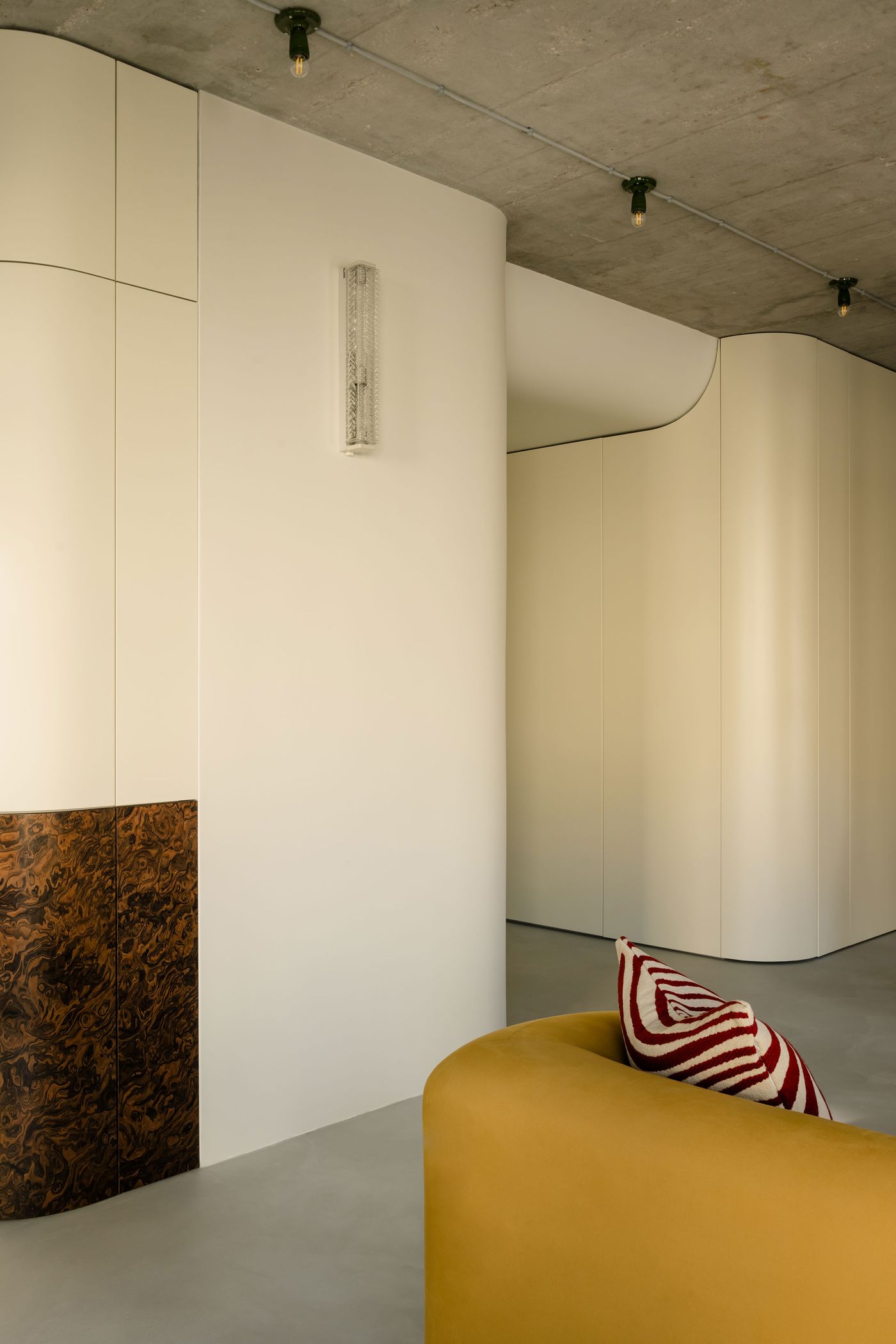 A space featuring smooth, curved walls in a creamy off-white, creating a sense of fluid movement with a richly veined veneer section on the lower left side providing a stark, organic contrast. Overhead, the raw concrete ceiling with exposed track lighting hints at an industrial influence. A vibrant mustard yellow sofa with a bold, striped throw pillow introduces a pop of color. A space featuring smooth, curved walls in a creamy off-white, creating a sense of fluid movement with a richly veined veneer section on the lower left side providing a stark, organic contrast. Overhead, the raw concrete ceiling with exposed track lighting hints at an industrial influence. A vibrant mustard yellow sofa with a bold, striped throw pillow introduces a pop of color.