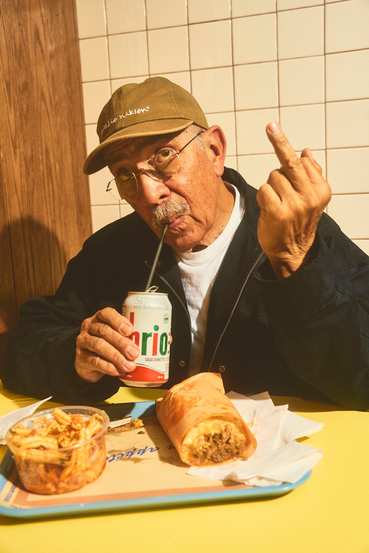 An older man wearing a cap and glasses sips a "BRIO" soda while holding a sandwich. He gestures with his middle finger, adding a touch of playful defiance. The scene is set in the retro-styled interior of Renzo Sandwich, with wood paneling and tiled walls visible. A side container of fries completes the casual meal.