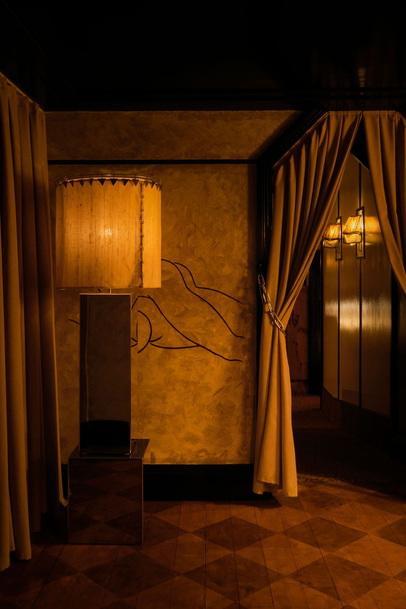 A luxurious, retro-inspired space in The Roses of Elagabalus, featuring a tall, grand floor lamp with a textured, circular shade and a mirrored base, casting a soft, warm light. The golden-brown wall is adorned with a delicate, abstract line drawing, while heavy, chain-held drapes frame an inviting, dimly lit passage. A luxurious, retro-inspired space in The Roses of Elagabalus, featuring a tall, grand floor lamp with a textured, circular shade and a mirrored base, casting a soft, warm light. The golden-brown wall is adorned with a delicate, abstract line drawing, while heavy, chain-held drapes frame an inviting, dimly lit passage.