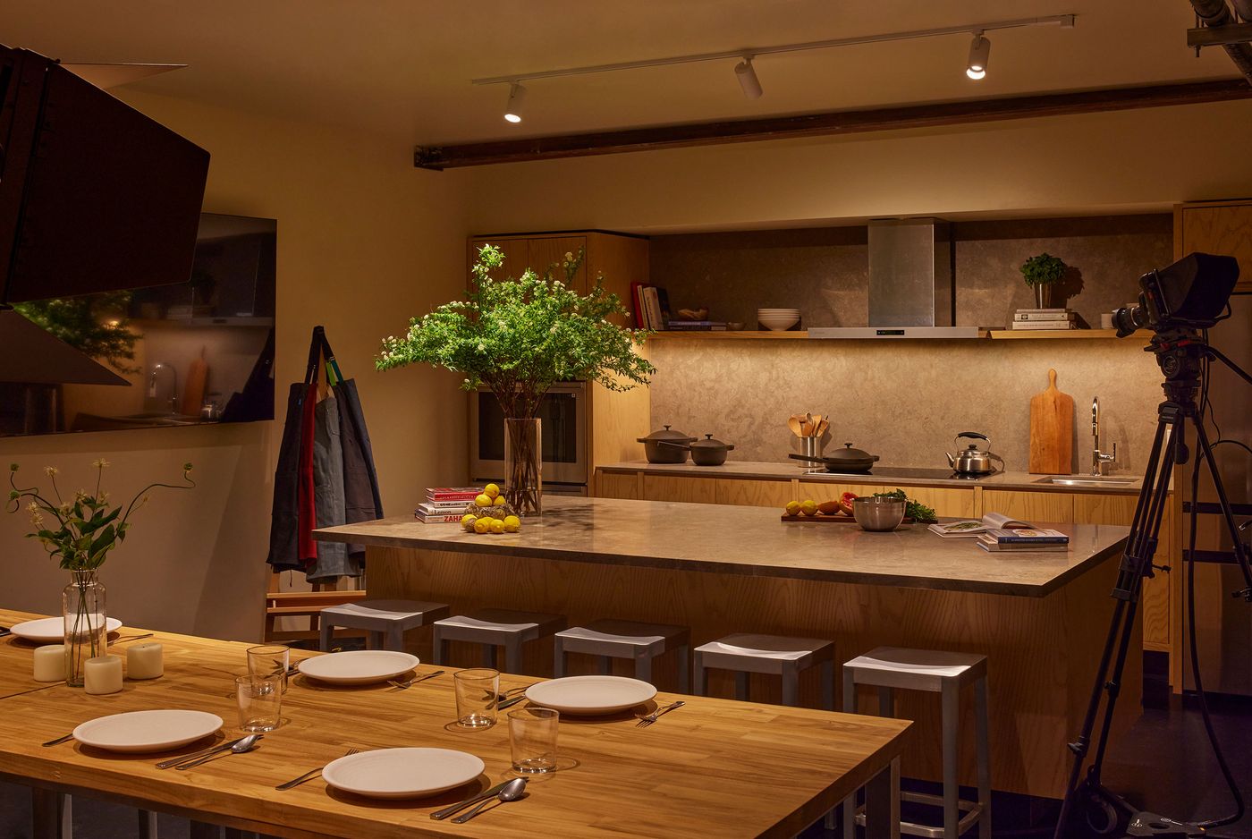 A warm, modern kitchen with a wooden dining table set, decorative plants, cookware, and a camera ready for filming. A warm, modern kitchen with a wooden dining table set, decorative plants, cookware, and a camera ready for filming.