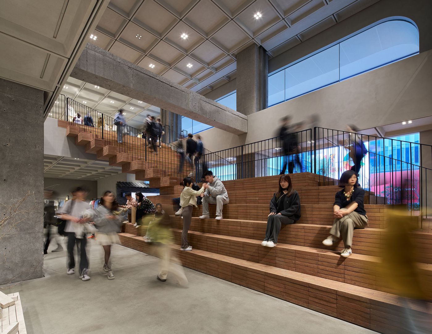 A bustling modern interior featuring people walking and sitting on brick steps, surrounded by concrete and large windows. A bustling modern interior featuring people walking and sitting on brick steps, surrounded by concrete and large windows.