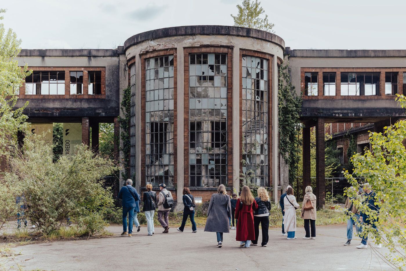 A group of people explore the overgrown courtyard of a derelict industrial building with a striking cylindrical glass façade, partially shattered, surrounded by decaying concrete and creeping vegetation. A group of people explore the overgrown courtyard of a derelict industrial building with a striking cylindrical glass façade, partially shattered, surrounded by decaying concrete and creeping vegetation.