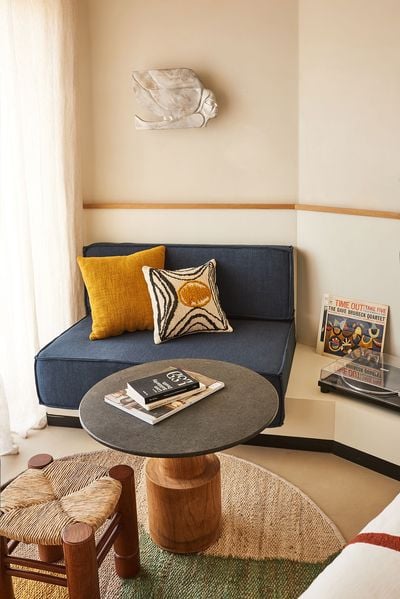 A cosy seating corner at Ace Hotel & Swim Club Athens combines a deep-blue upholstered sofa, mustard and patterned cushions, and a round stone-topped table with a sculptural timber base. A woven stool, circular natural-fibre rug, vintage record player and Dave Brubeck album add warmth and retro character, complemented by a carved wall relief and softly filtered daylight. A cosy seating corner at Ace Hotel & Swim Club Athens combines a deep-blue upholstered sofa, mustard and patterned cushions, and a round stone-topped table with a sculptural timber base. A woven stool, circular natural-fibre rug, vintage record player and Dave Brubeck album add warmth and retro character, complemented by a carved wall relief and softly filtered daylight.
