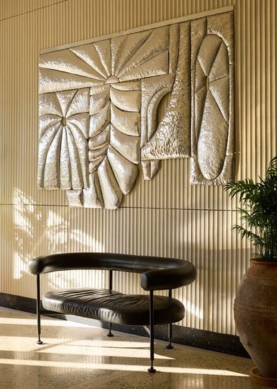 A sculptural metallic wall relief with abstract geometric and botanical forms dominates a sunlit interior at Ace Hotel & Swim Club Athens. Mounted against vertically ribbed cream panelling, the textured artwork hangs above a curved black leather bench with slender metal legs. A large terracotta planter introduces greenery, reinforcing the interplay of modernist design and Mediterranean references. A sculptural metallic wall relief with abstract geometric and botanical forms dominates a sunlit interior at Ace Hotel & Swim Club Athens. Mounted against vertically ribbed cream panelling, the textured artwork hangs above a curved black leather bench with slender metal legs. A large terracotta planter introduces greenery, reinforcing the interplay of modernist design and Mediterranean references.