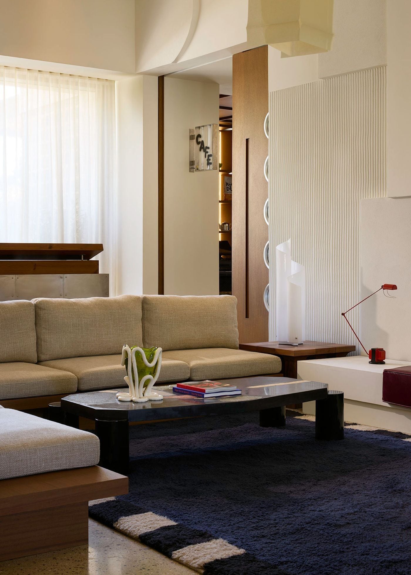 A contemporary lounge at Ace Hotel & Swim Club Athens combines cream-coloured modular sofas, a low black coffee table and a plush navy rug with bold geometric borders. Ribbed wall panels, sculptural lighting, timber partitions and a red adjustable lamp lend the space a playful modernist character, while daylight filters through floor-to-ceiling curtains.