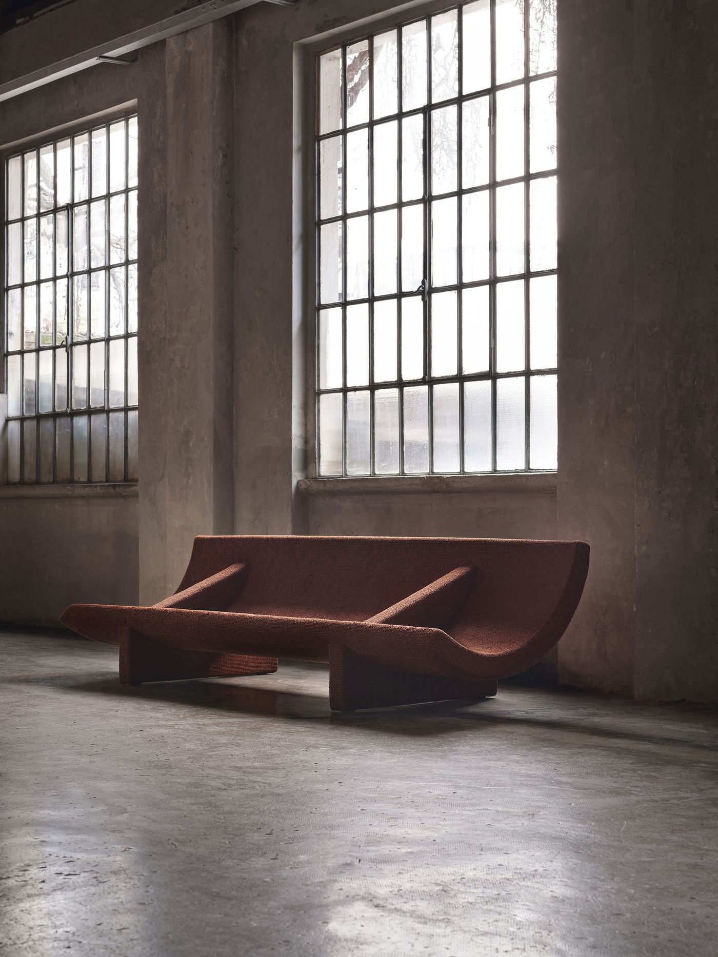 Sculptural deep-red sofa with angular armrests, placed in a raw industrial space with large factory-style windows. Sculptural deep-red sofa with angular armrests, placed in a raw industrial space with large factory-style windows.